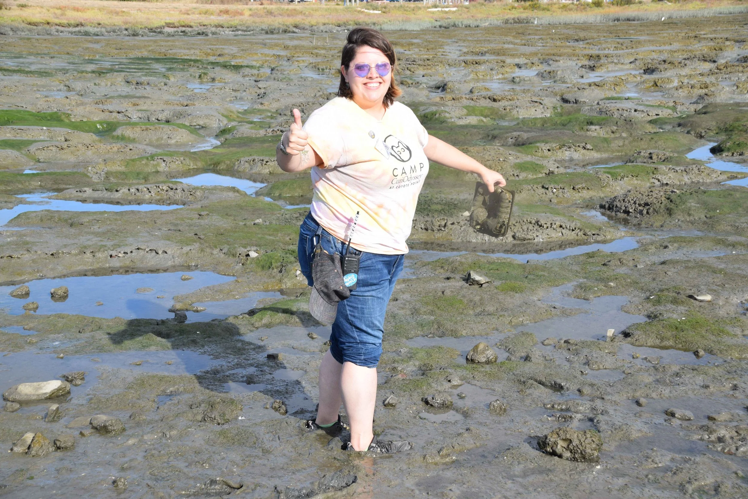 A photo of a person standing ankle-deep in a muddy marsh. They are white with brown hair, and they are wearing a t-shirt and jeans rolled up to the knees. They're giving a big thumbs-up and holding a piece of litter they pulled out of the mud.