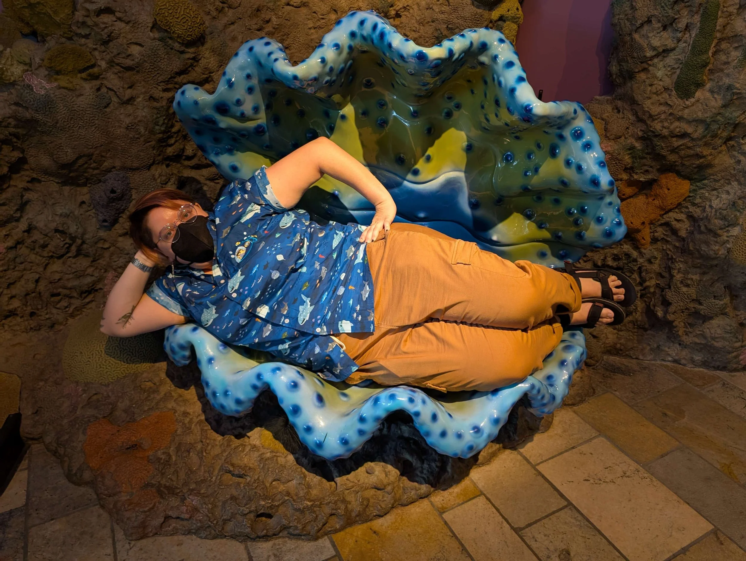 Emma lying on their side in a giant scallop shell sculpture. Emma is wearing a blue shirt pattenred with sharks, tan pants, and black sandals.