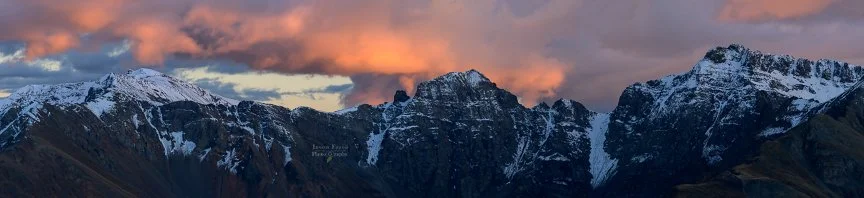 Snow-covered San Juan Mountains beneath glowing orange sunset clouds viewed from the top of Corkscrew Pass, Colorado.