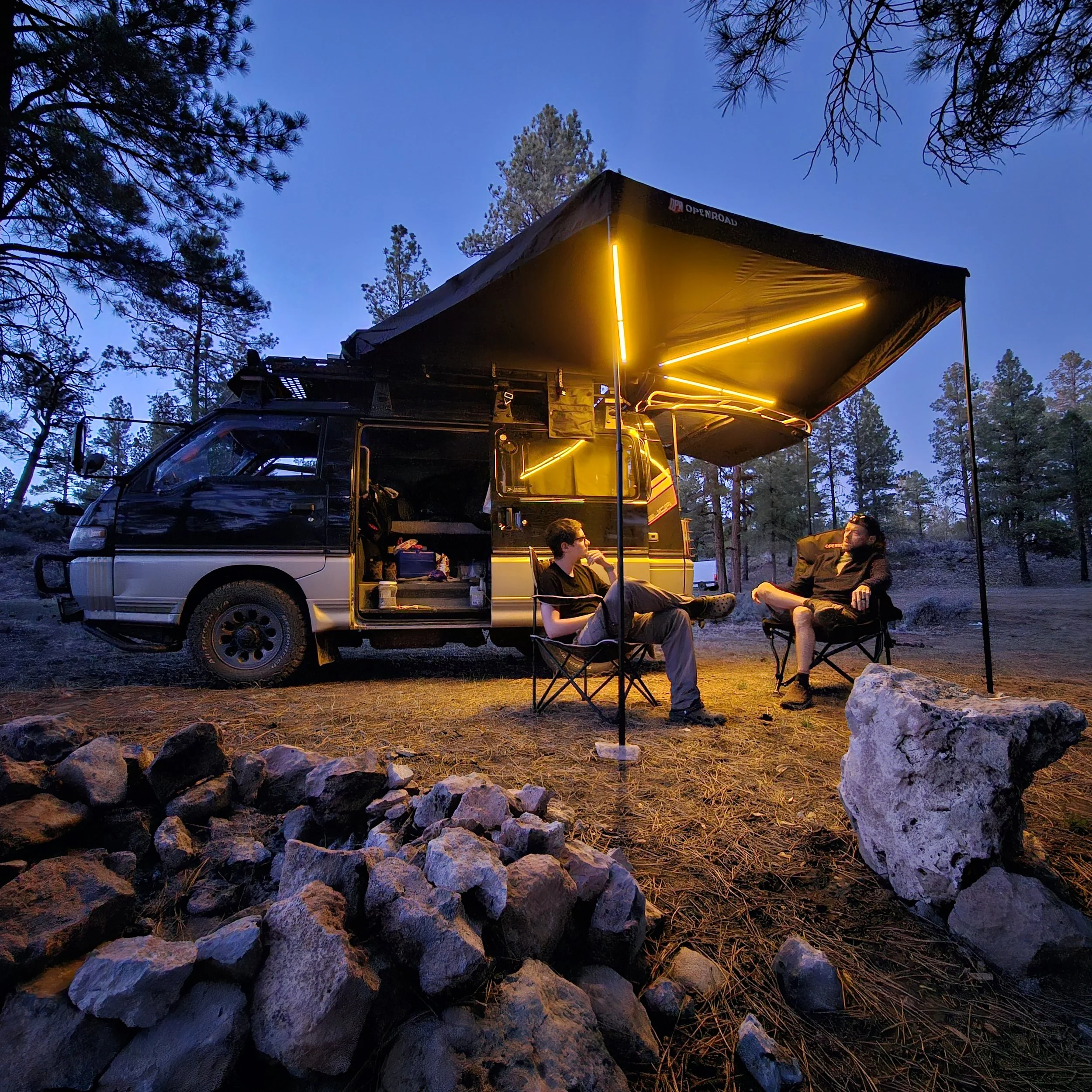 Two people sitting beside a Mitsubishi Delica L300 beneath an OPENROAD 270-degree awning illuminated by its integrated LED lights.