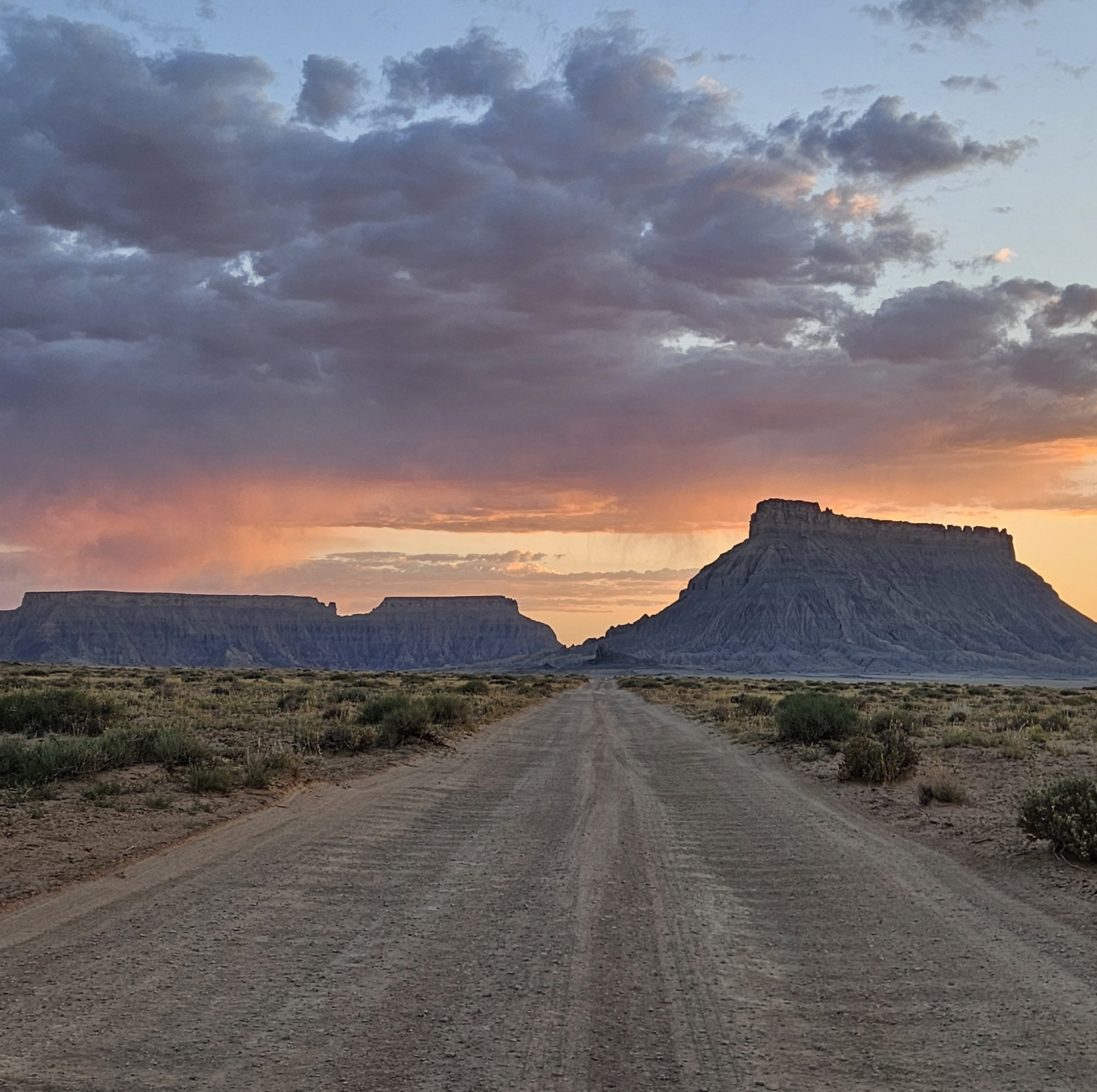 Looking down a straight desert dirt road off toward Factory Butte and the day's sunset sky full of oranges, pinks, and purples.