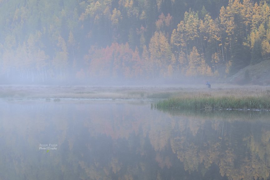 A lone photographer stands in a foggy meadow beside Crystal Lake, Colorado, surrounded by golden autumn aspens and soft reflections in the still water.