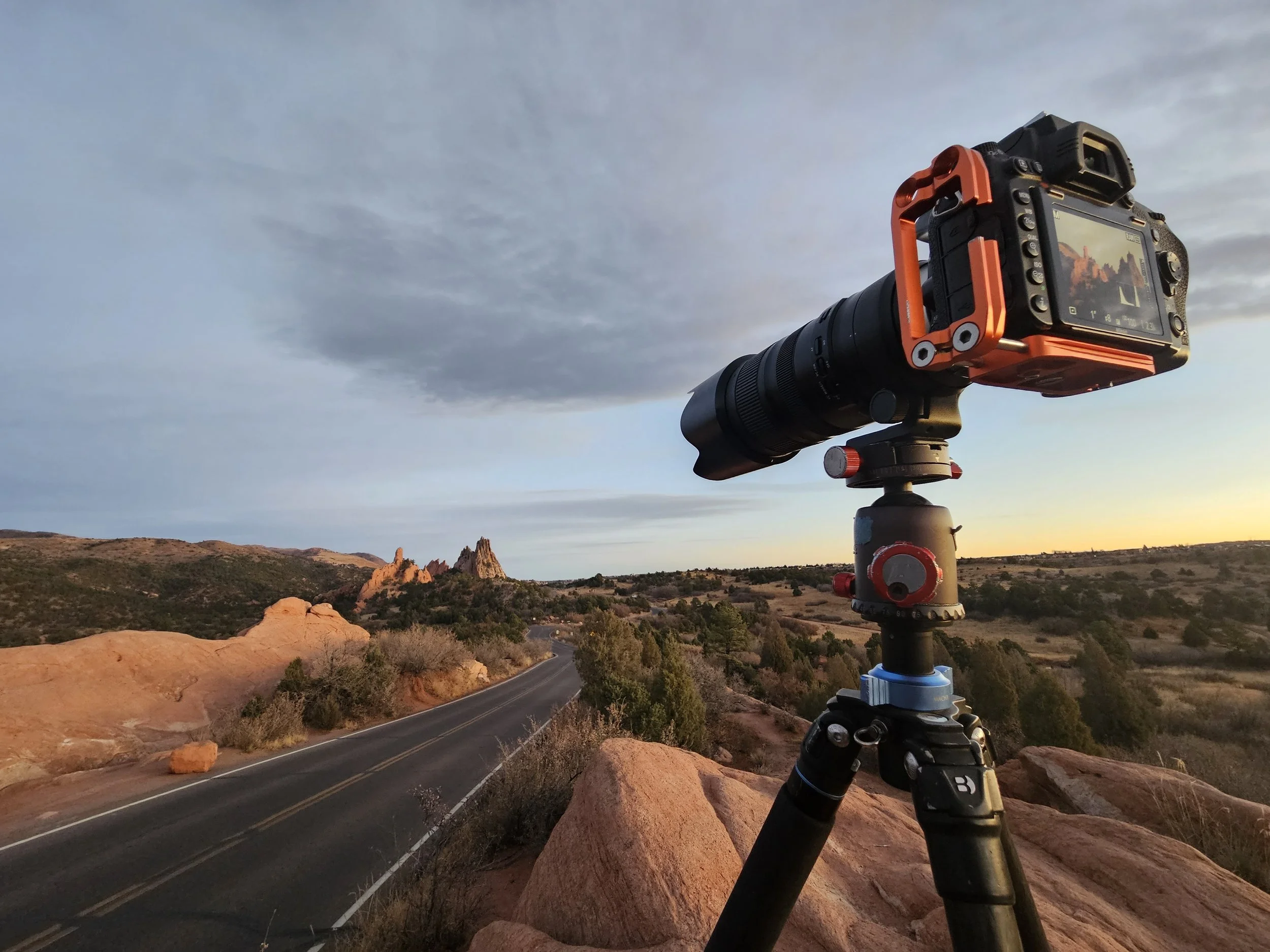 A camera mounted on a tripod in a desert landscape with a winding road, rocks, hills, and a cloudy sky at sunset.