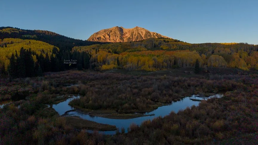 Aerial drone photograph of sunrise illuminating a mountain peak above golden autumn aspens and a winding creek near Kebler Pass, Colorado.