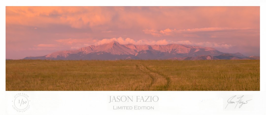 An old two track trail leads off into the distance as it travels through a field of tall gras and leads the way toward Pikes Peak standing tall in the predawn sunlight.