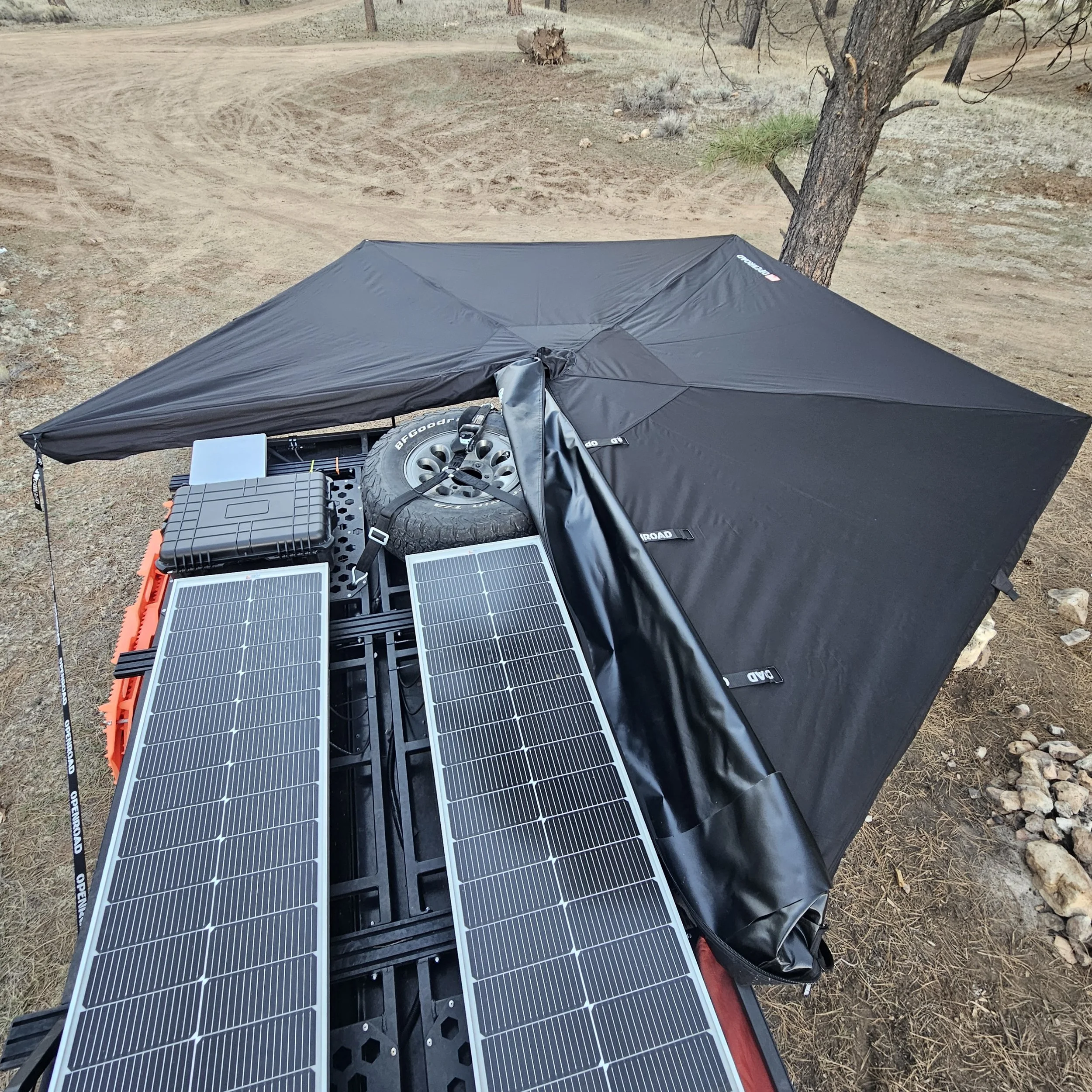 Overhead view of an OPENROAD 270-degree awning deployed around a Mitsubishi Delica L300 roof rack with solar panels and a spare tire.
