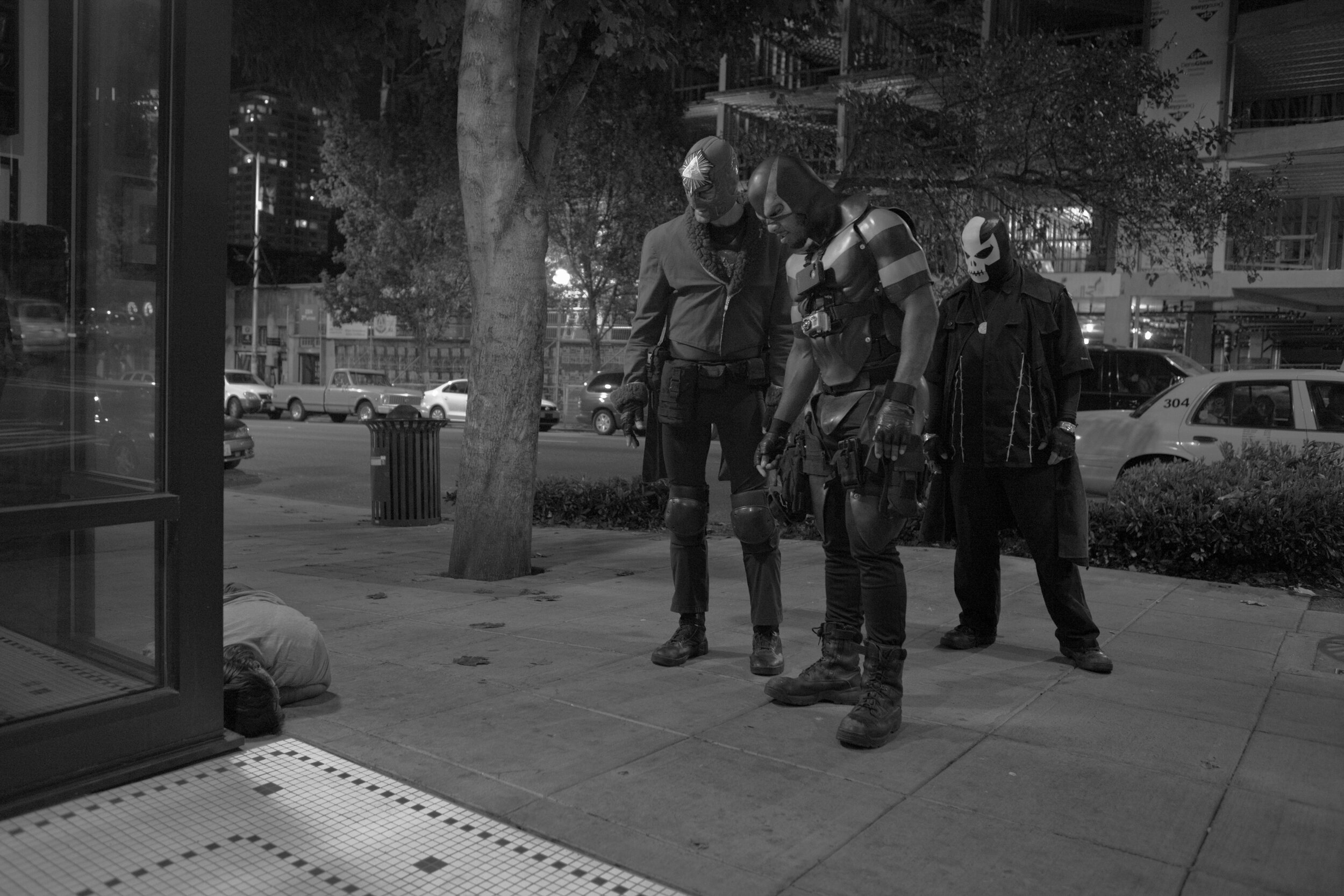  El Caballero, Phoenix Jones, and Westlake Drake look at a drunk man passed out on the street in Seattle, WA on Oct. 6, 2012. 
