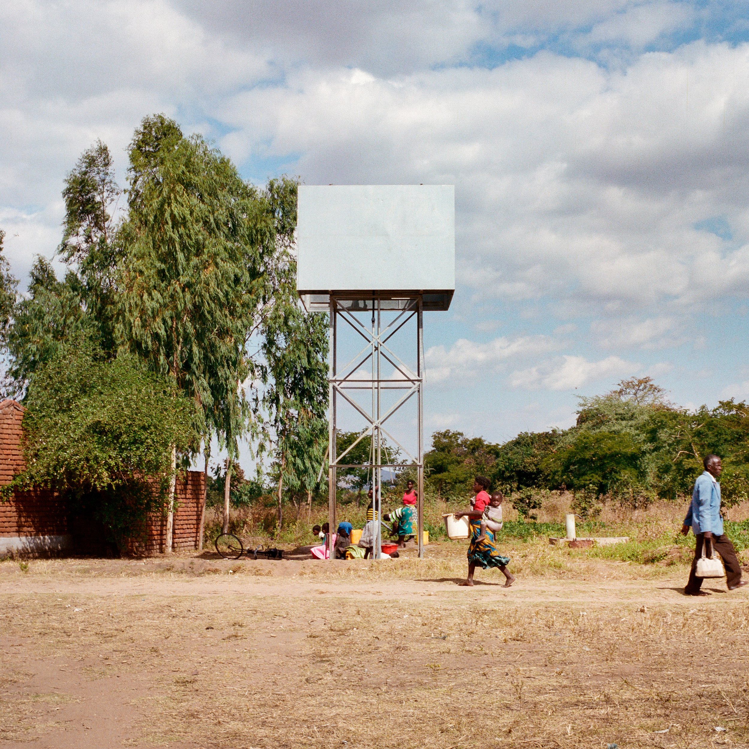  Empty, unused water tower from the site of a broken PlayPump. Malopa Primary School in Mora, Balaka District, Malawi. Photographed June 13, 2015. The people getting water underneath the tower are using a handpump that does not require use of the tow