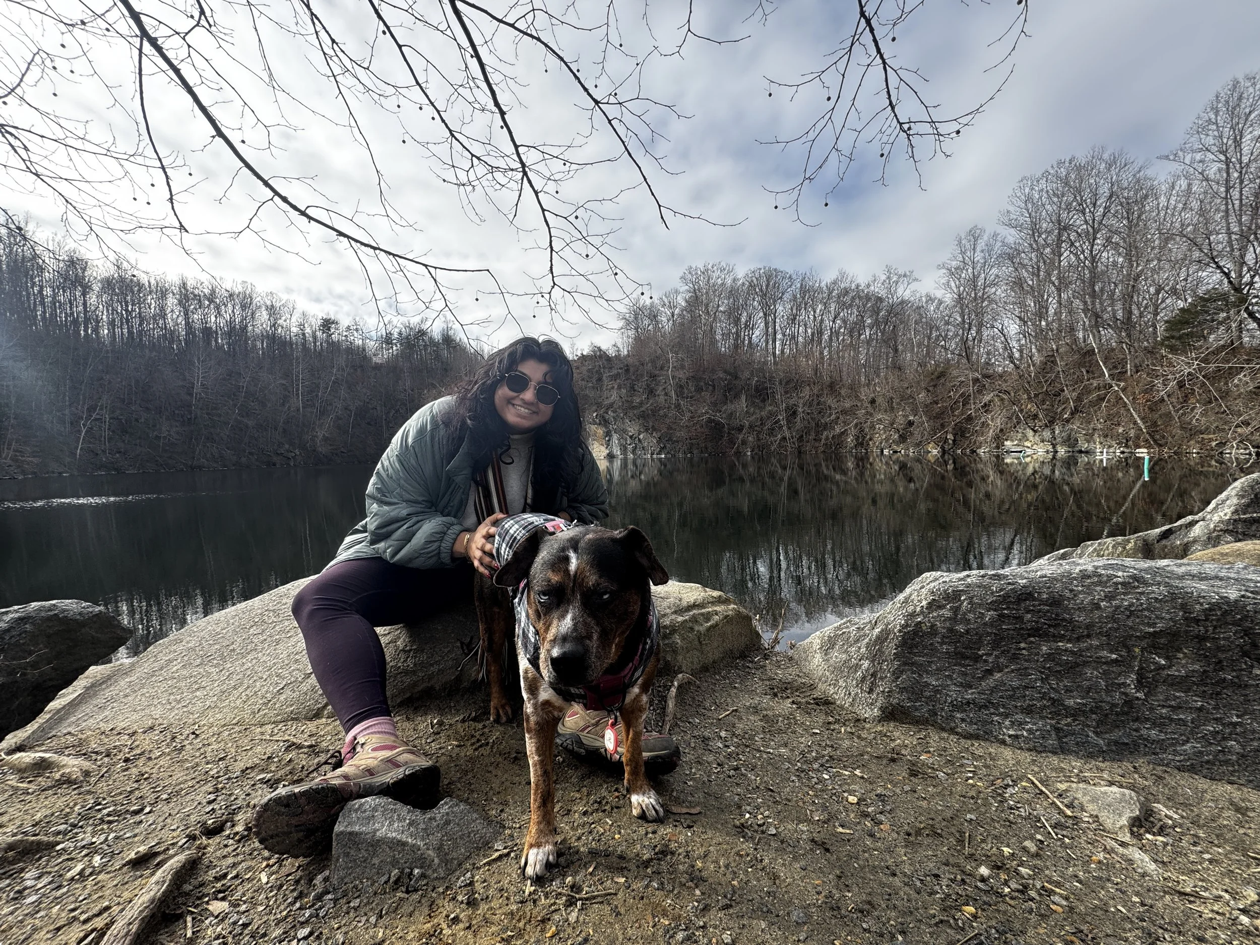 A woman and her dog by a lake in a wooded area during winter. The woman is sitting on a rock, smiling, wearing sunglasses, a jacket, and hiking shoes. The dog, a black and brown mixed breed, is standing in front of her, wearing a harness. The background shows leafless trees and cloudy sky reflected on the calm lake.