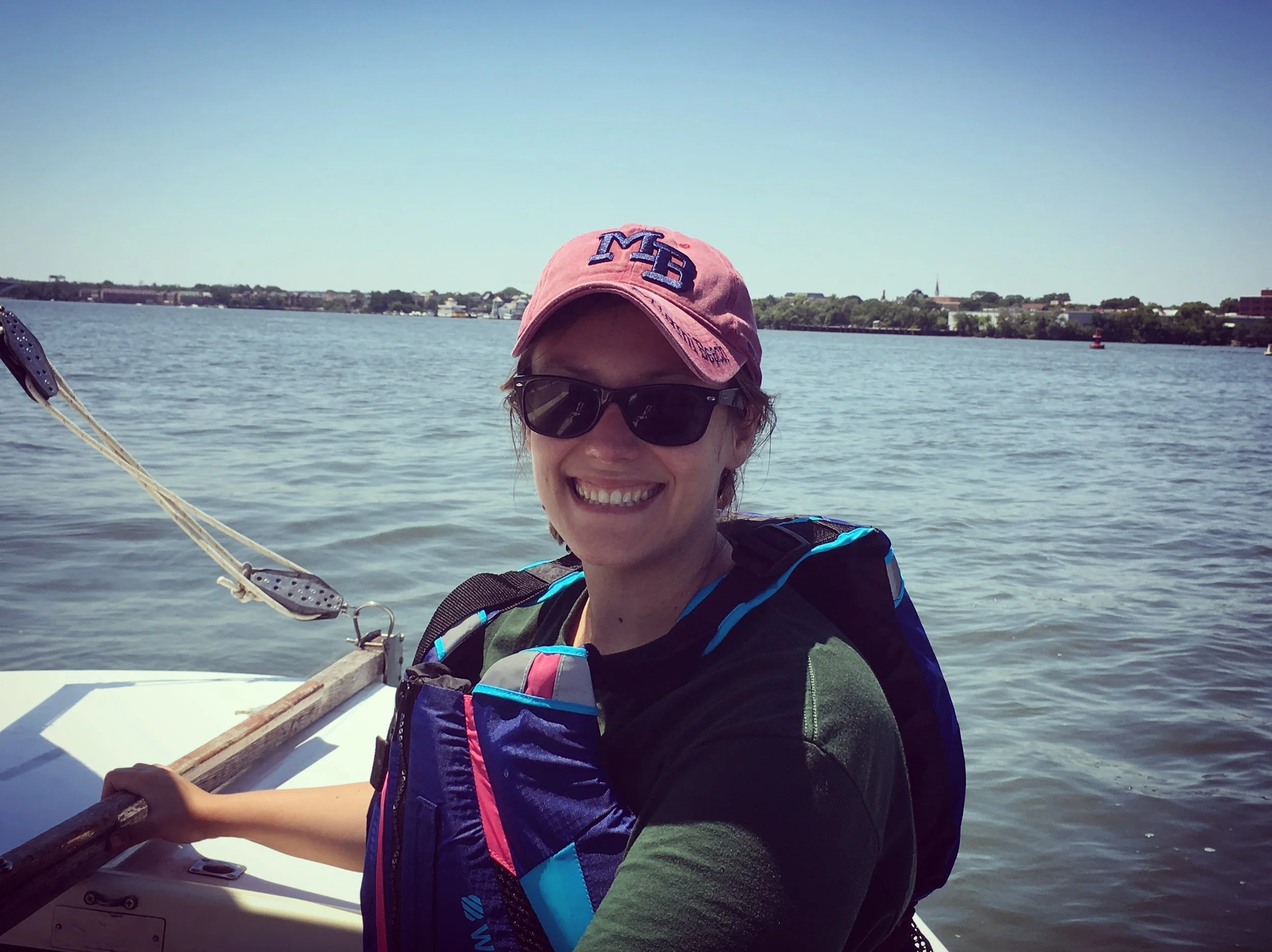 A smiling woman wearing sunglasses and a pink cap, sitting in a boat on a body of water with a distant shoreline and blue sky in the background.