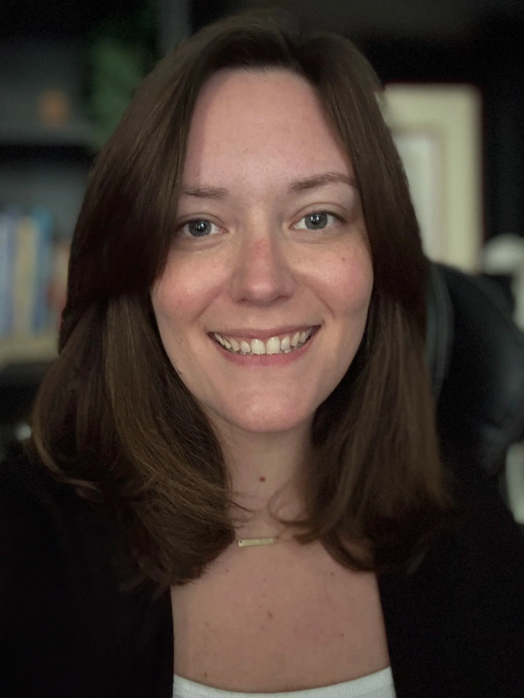 Leslee Bry is smiling warmly. She has long brown hair, freckles, blue eyes, wearing a white top and a necklace with a horizontal gold bar, in a dark room with blurred shelves in the background.