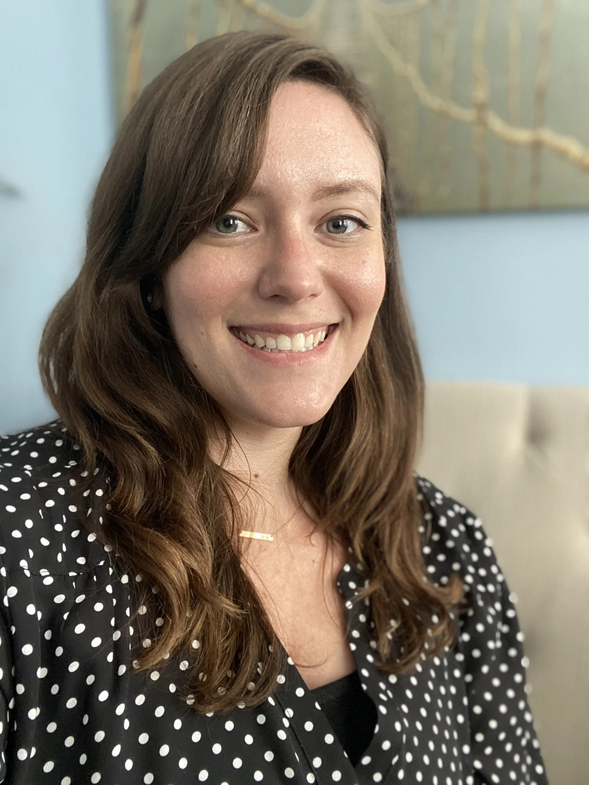 A woman with wavy brown hair and blue eyes smiling, wearing a black polka dot blouse and a gold necklace, sitting in front of a blue wall with a painting.