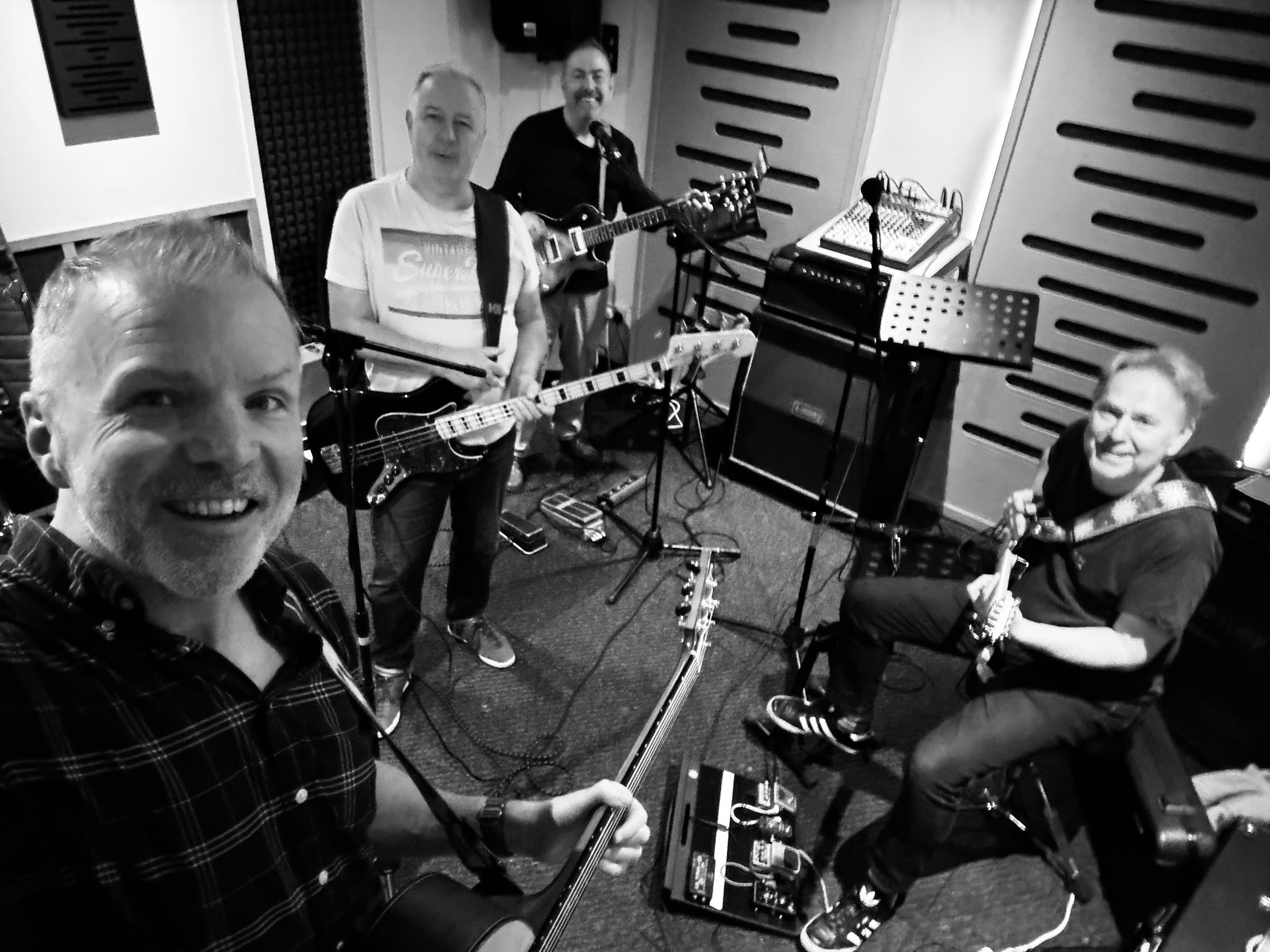 Five people in a music studio, four playing guitars and one taking a selfie, surrounded by musical equipment.