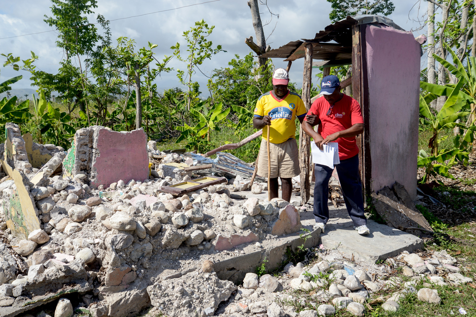 A CBM staff member helps a man walk through the rubble with his cane