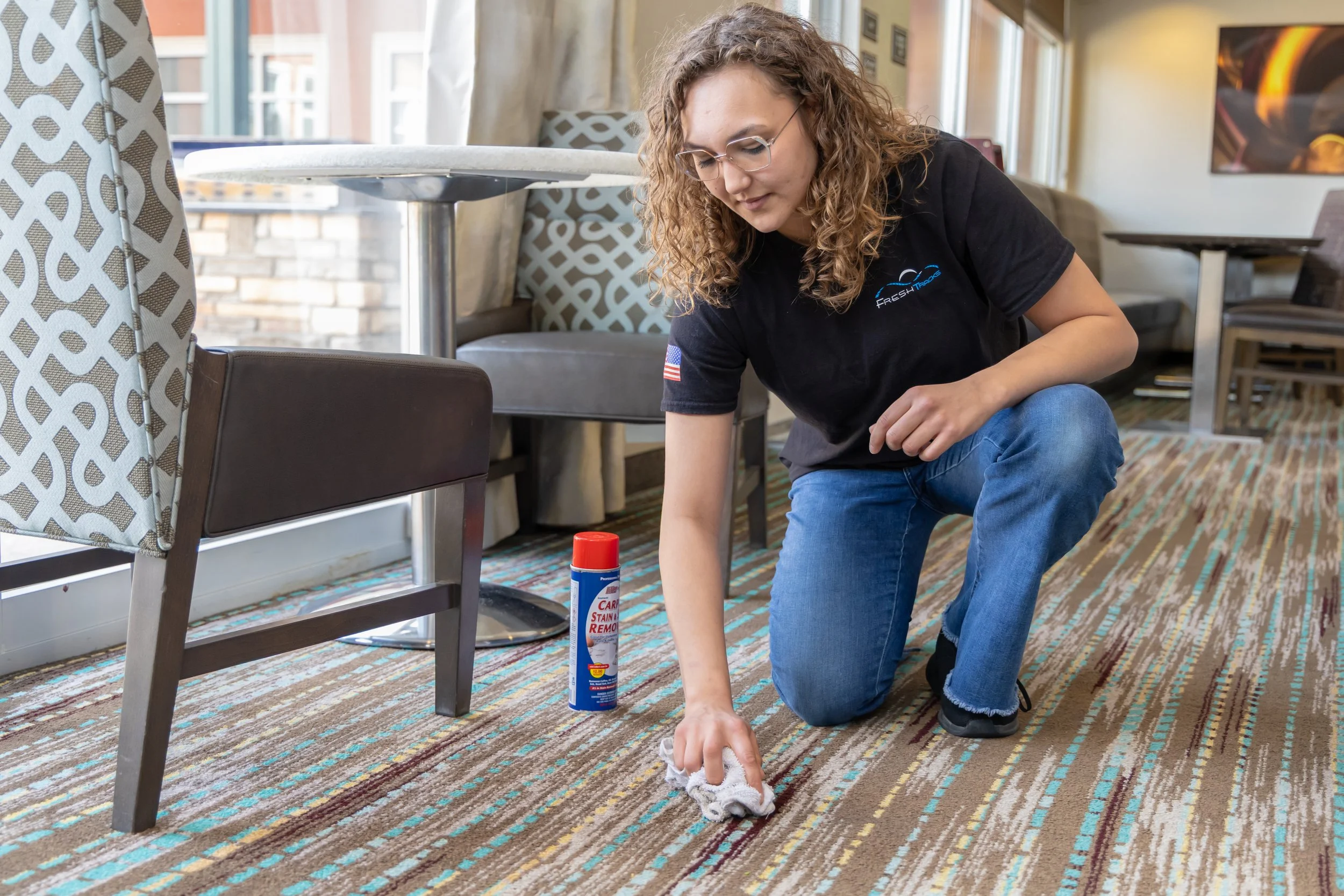 A Fresh Tracks team member kneeling on the floor and using a cloth with carpet stain remover to spot clean a stain on patterned carpet in a hotel lobby lounge area, with modern seating and large windows visible in the background.