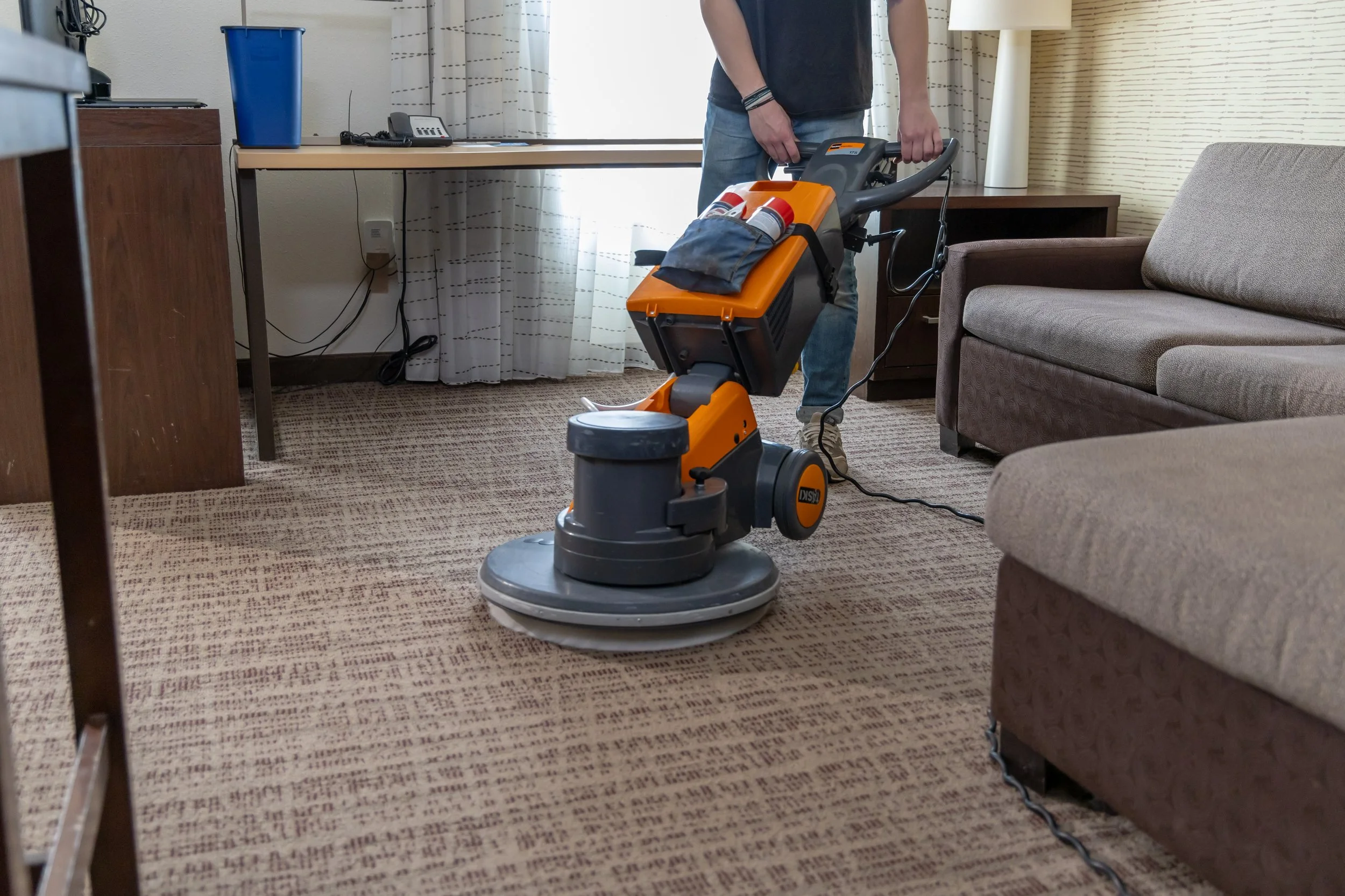 A Fresh Tracks staff member operating a professional orange and gray TASKI floor cleaning machine on patterned carpet inside a guest room, with a sofa, desk, and window area visible in the background.