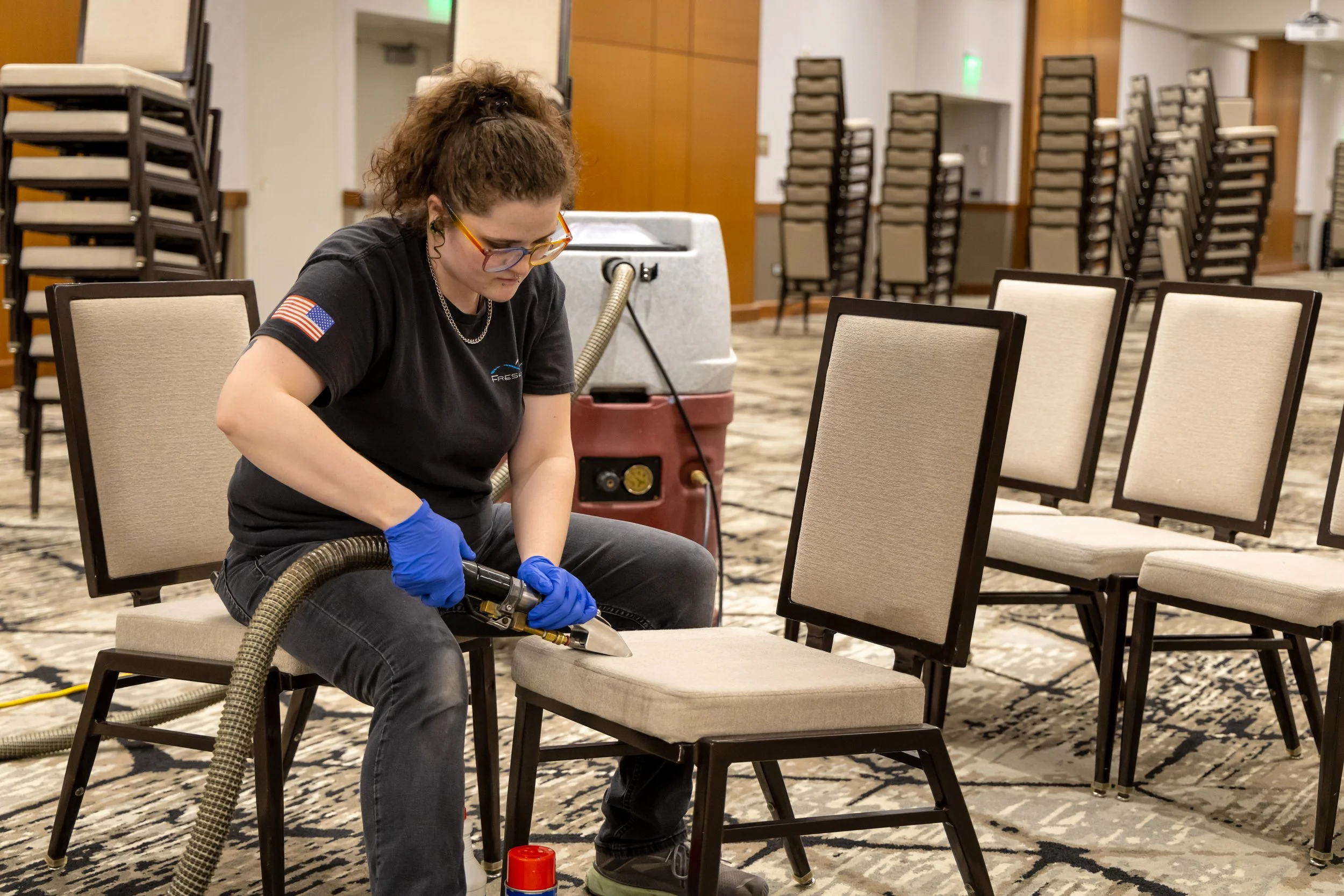 A Fresh Tracks team member using professional upholstery cleaning equipment to clean the seat of a chair in a large event center, with rows of chairs and cleaning supplies visible around her.