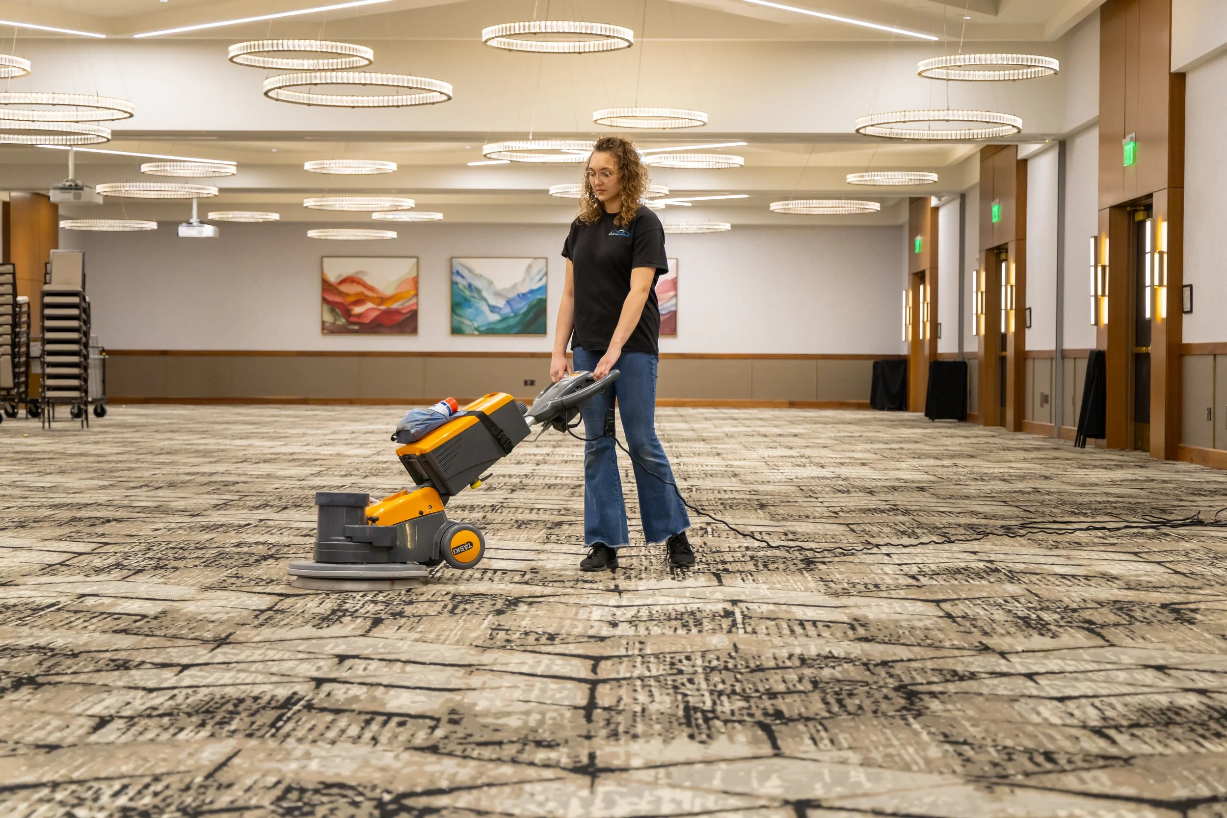 A Fresh Tracks team member operating a professional orange and gray TASKI floor cleaning machine on patterned carpet in a large, modern event center ballroom, with contemporary lighting and artwork visible in the background.