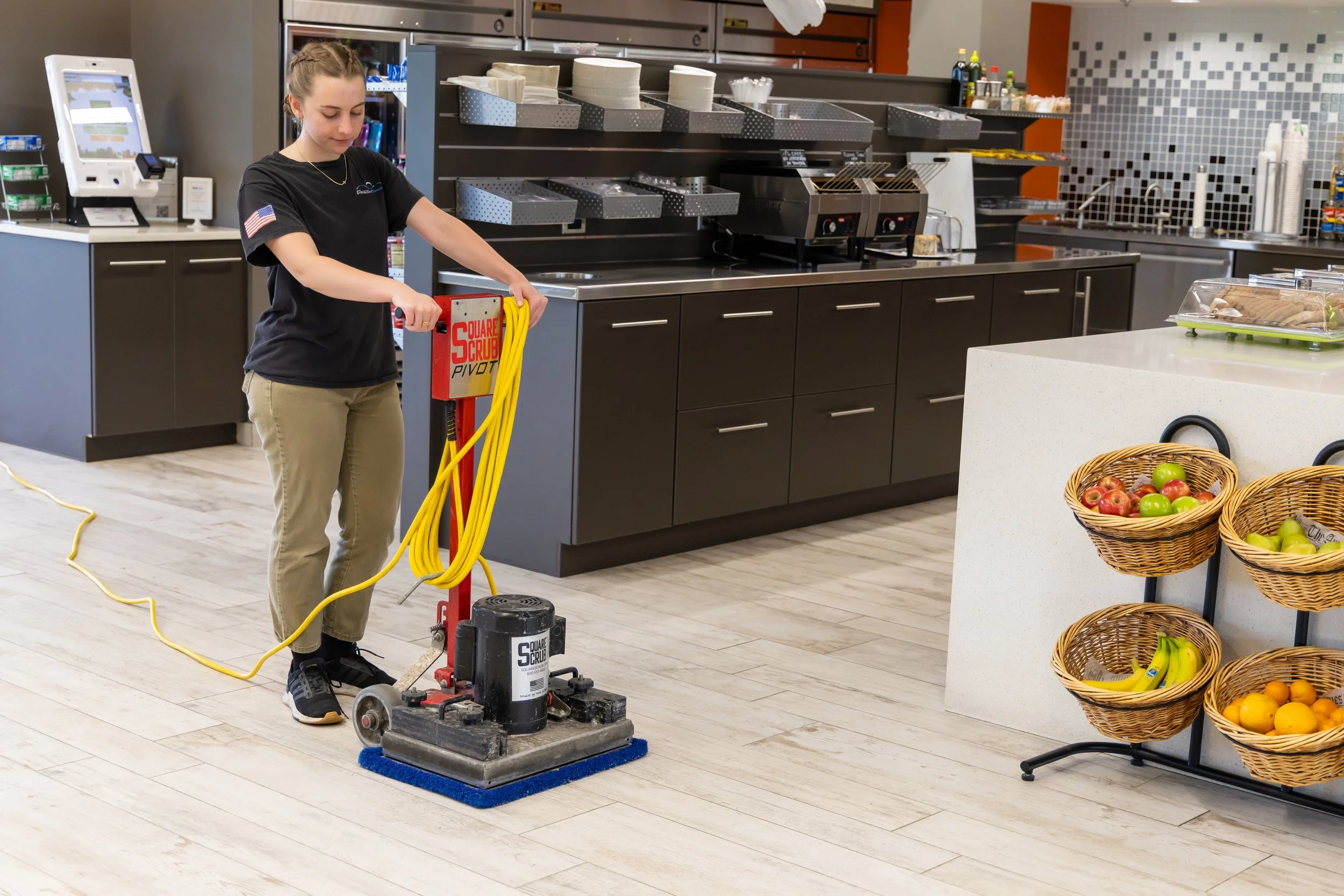 Fresh Tracks team member operating a Square Scrub floor cleaning machine in a modern hotel breakfast area with fresh fruit baskets and stainless steel counters