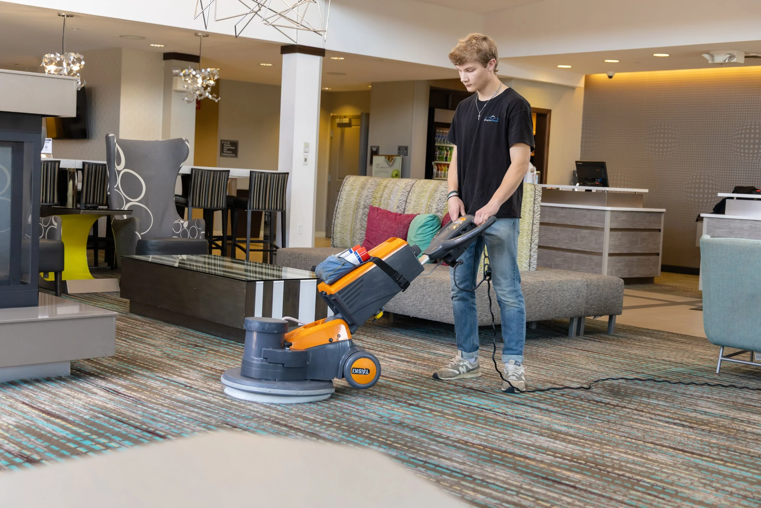 A Fresh Tracks team member operating a professional orange and gray TASKI floor cleaning machine on a patterned carpet in a modern hotel lobby lounge, with contemporary seating, a reception desk, and dining area visible in the background.