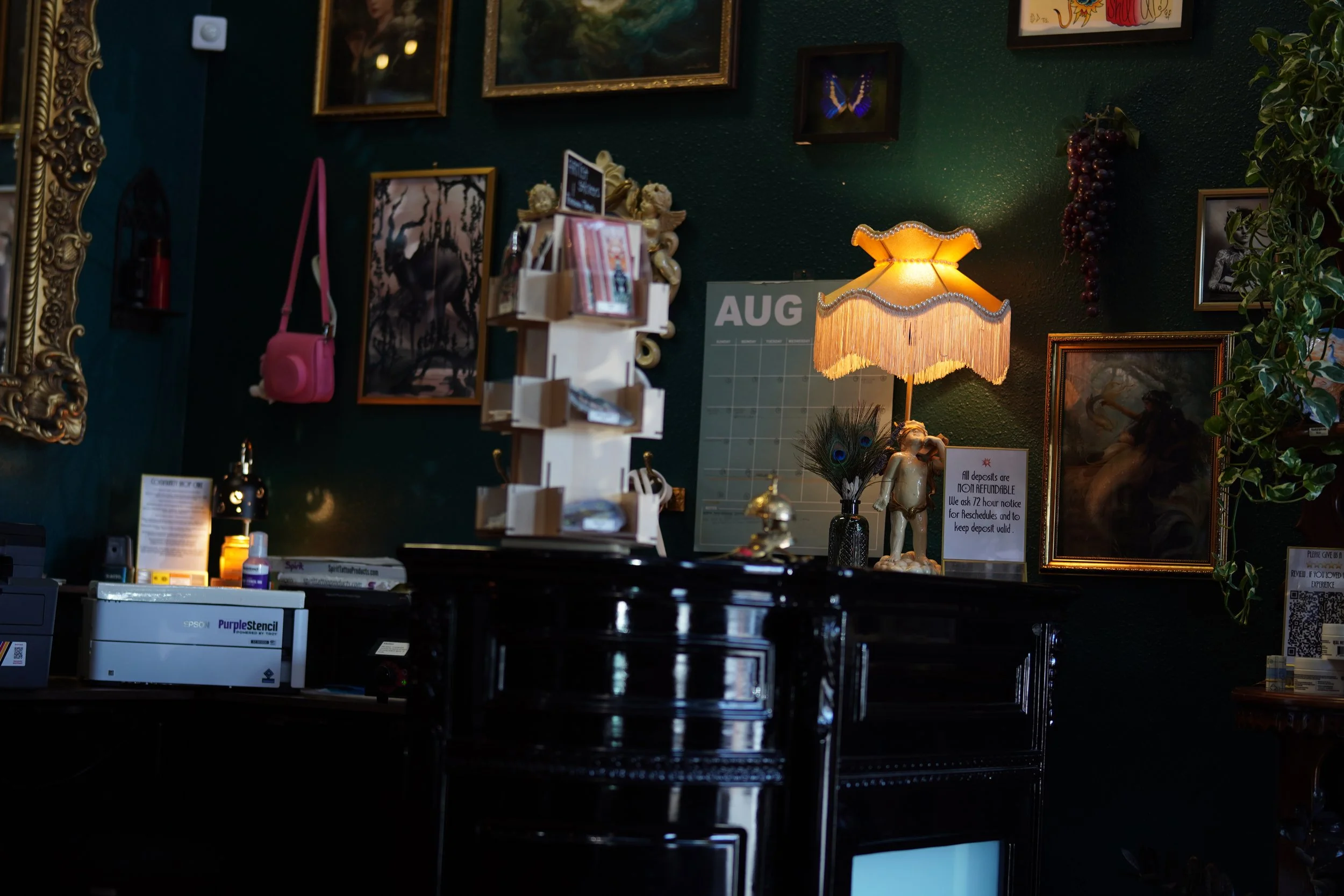 A dimly lit indoor space with framed artwork and decorations on a dark green wall, including a small pink bag, a vintage-style lamp with fringes, a decorative sculpture of a woman, a bunch of grapes, a peacock feather, and a calendar for August.