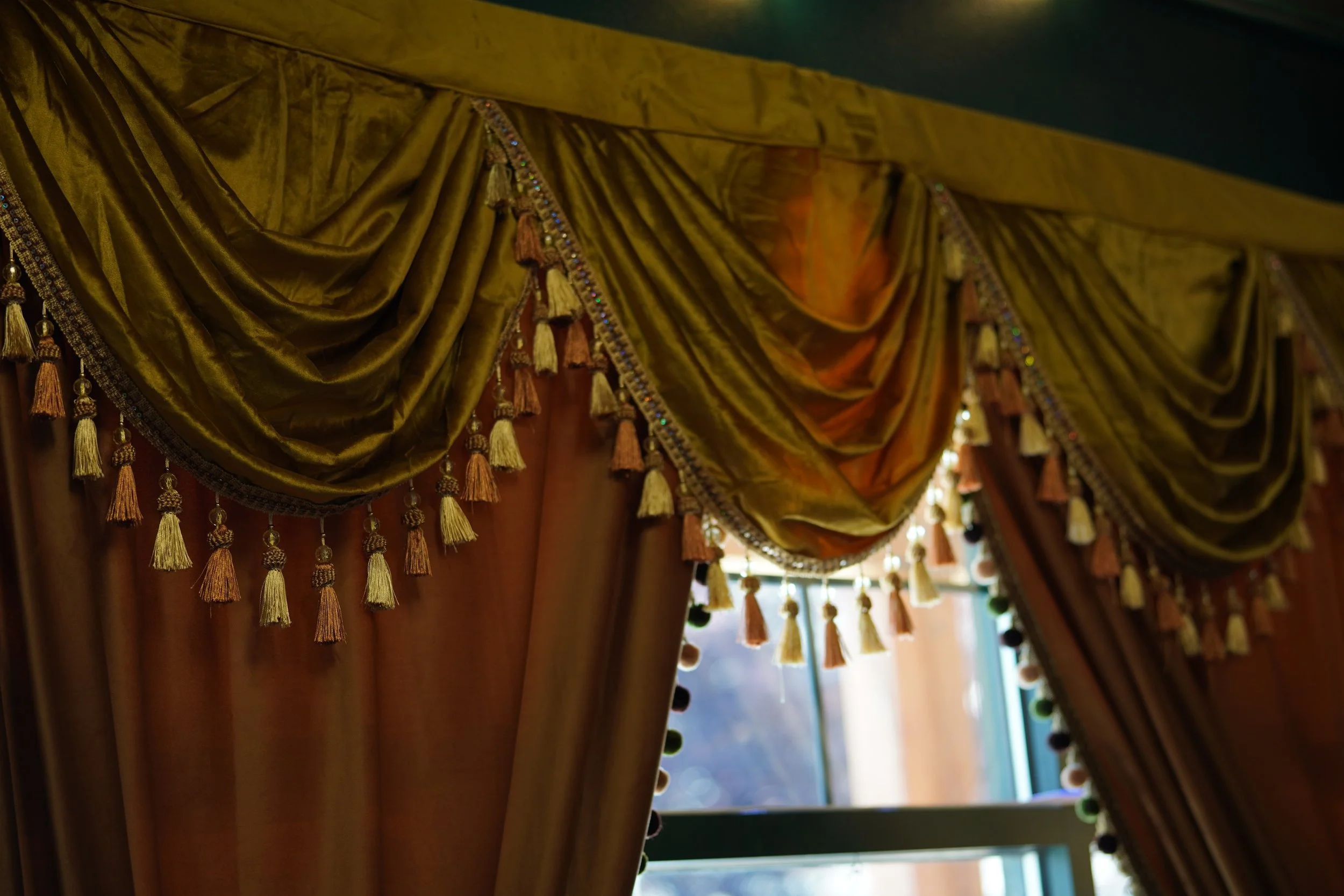 Decorative curtains with tassels and sequins hanging in front of a window with sunlight streaming through.