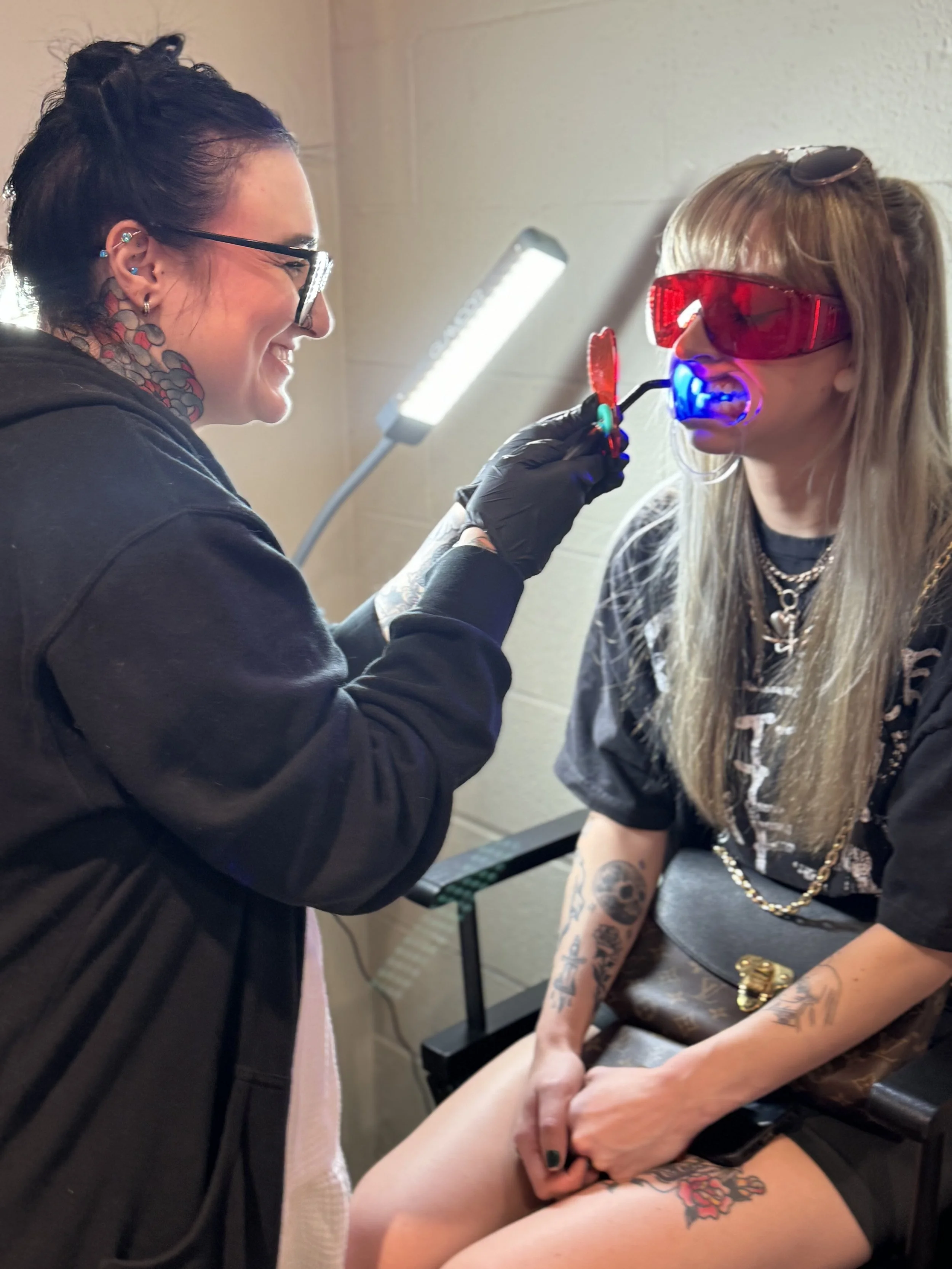 A tattoo artist with glasses, tattoos, and a black hoodie is tattooing a woman with long blonde hair, sunglasses, and tattoos on her arms and thigh. The woman is sitting on a chair, wearing a black T-shirt and shorts, holding a phone, while the artist is smiling and working on her thigh.