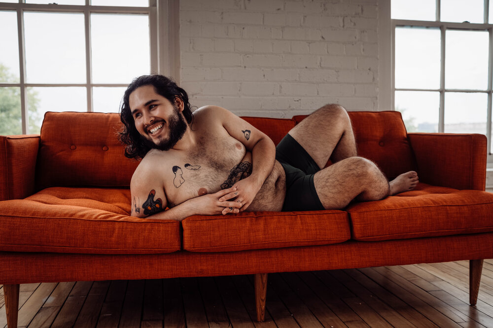 Oklahoma City Male Boudoir bearded shirtless man with tattoos lying on orange couch  and laughing