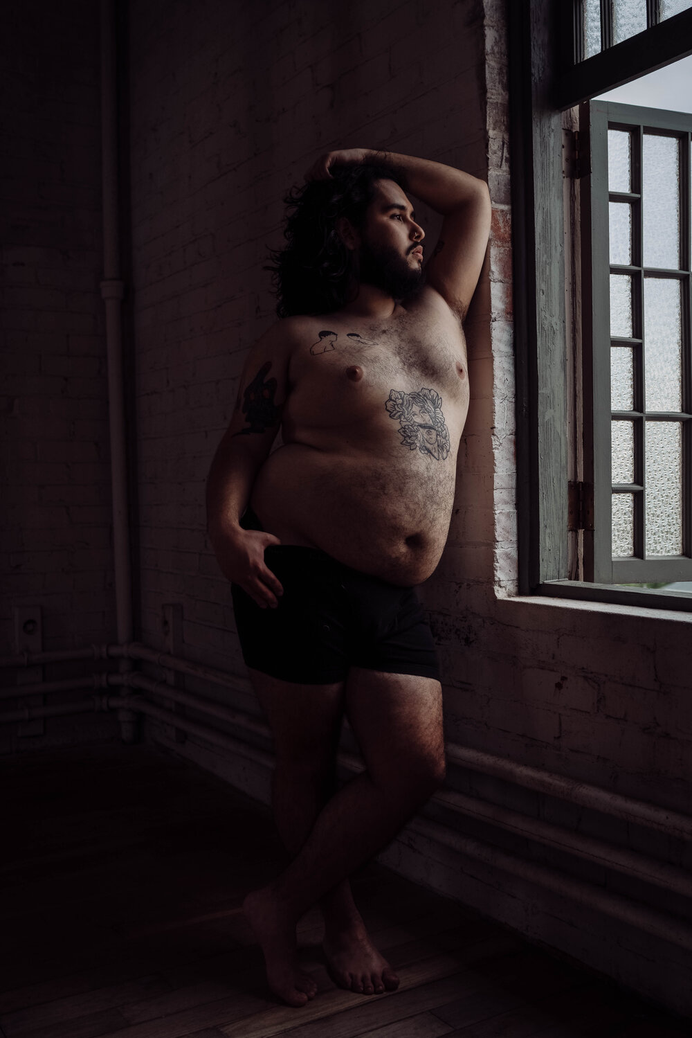 Oklahoma City Male Boudoir bearded shirtless man with tattoos leaning against brick wall looking out the window