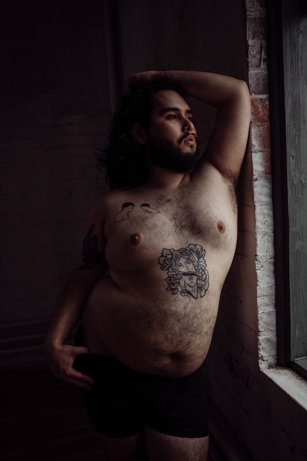 Oklahoma City Boudoir Photographer for Men bearded shirtless man with tattoos leaning against brick wall looking out the window