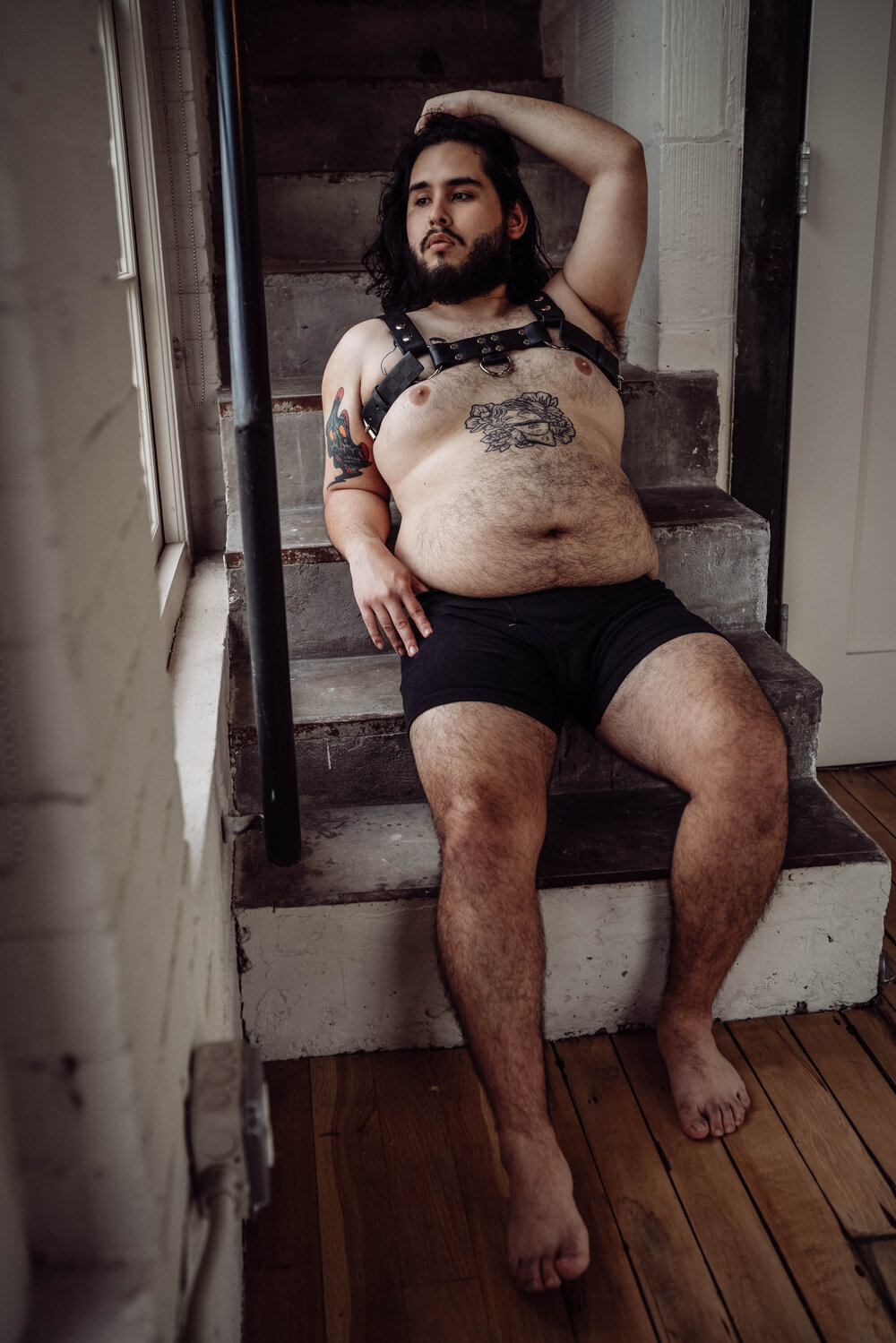 Houston Male Boudoir Photographer bearded man shirtless wearing a harness sitting on stairs