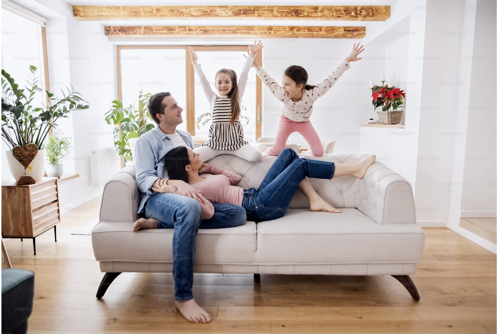 Family of four having fun on a beige couch in a bright living room with wooden flooring and large windows. Two young girls are playfully jumping on the couch, while the parents sit and relax nearby, smiling and enjoying the moment. The room is decorated with green plants and a poinsettia plant on a shelf.