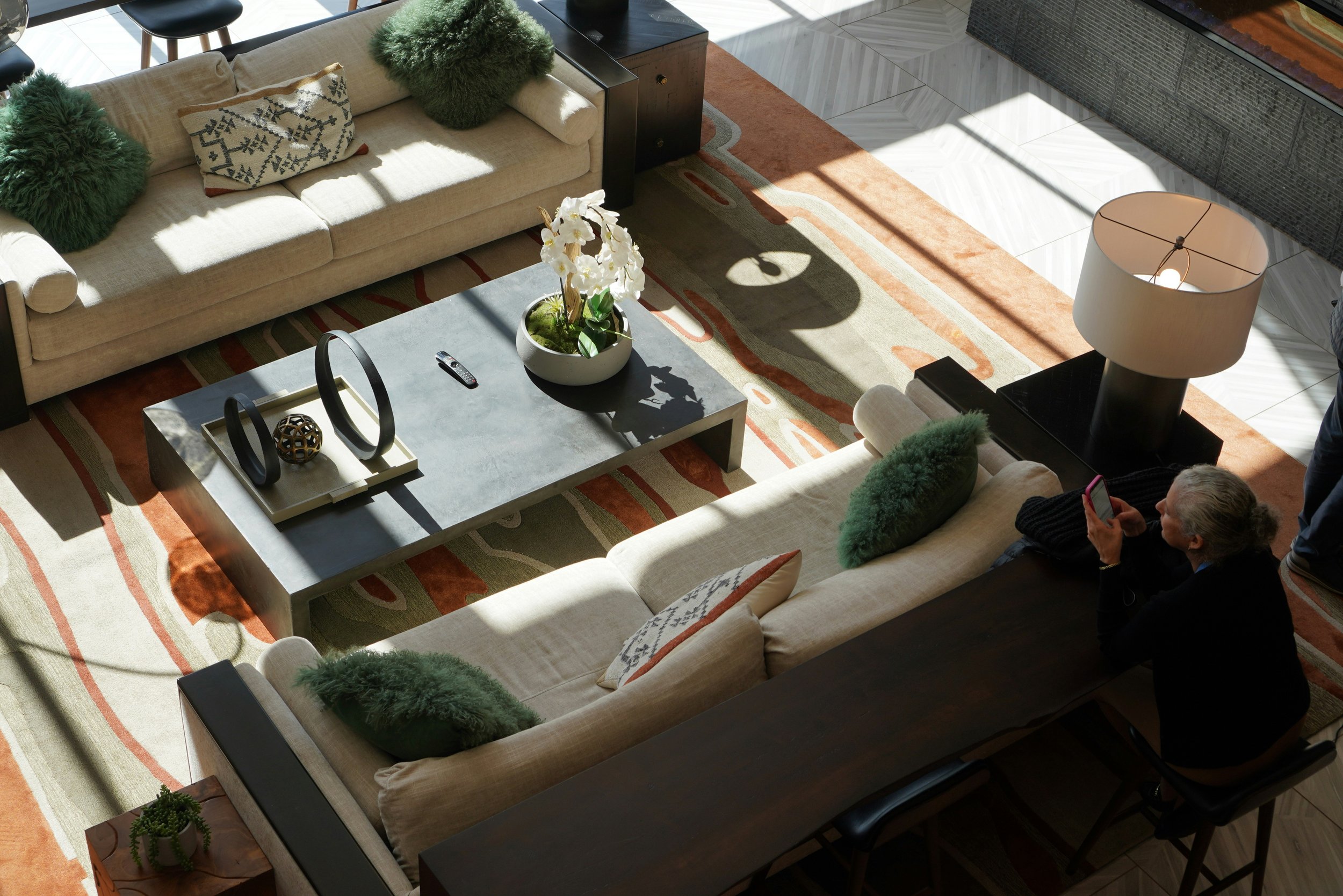 A cozy living room with a beige sofa decorated with green pillows and a patterned pillow. There is a light-colored coffee table with a potted white orchid, a remote control, and decorative objects. A large orange and beige patterned rug covers the floor. Sunlight streams through windows, casting shadows. A person and a woman are sitting on the sofa, and another person is standing nearby.