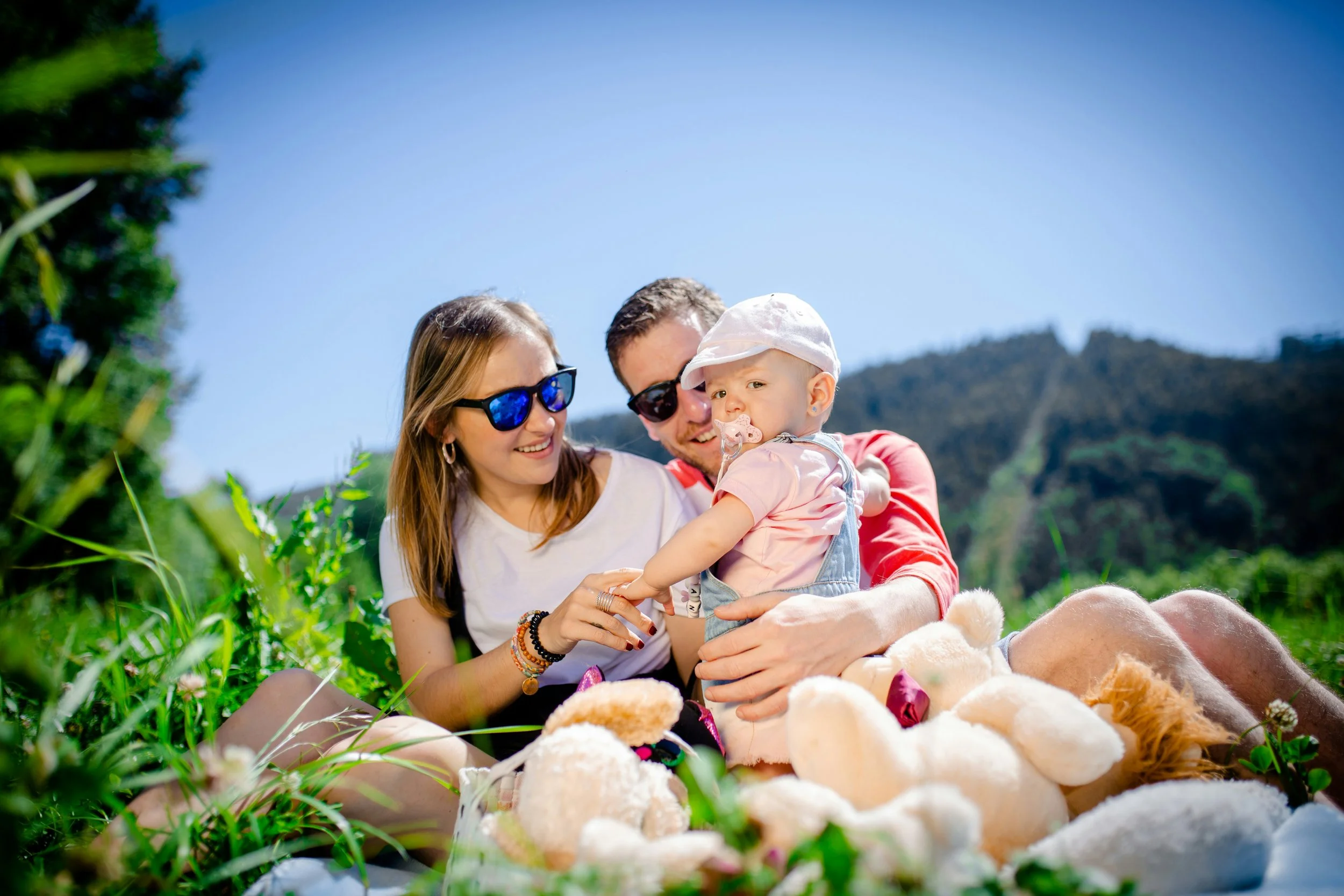 A family of three sitting together outdoors surrounded by toys, with a mountain in the background and clear blue sky, enjoying a sunny day.