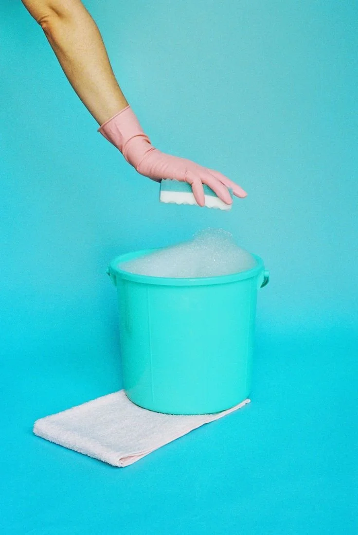 A hand wearing a pink rubber glove is scrubbing a bucket with a brush, placed on a towel against a blue background.