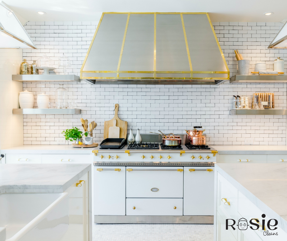 Bright white kitchen with a white oven and gold accents, white tile backsplash, open shelving with jars and dishes, cooking pots on the stove, cutting boards, and a potted plant.