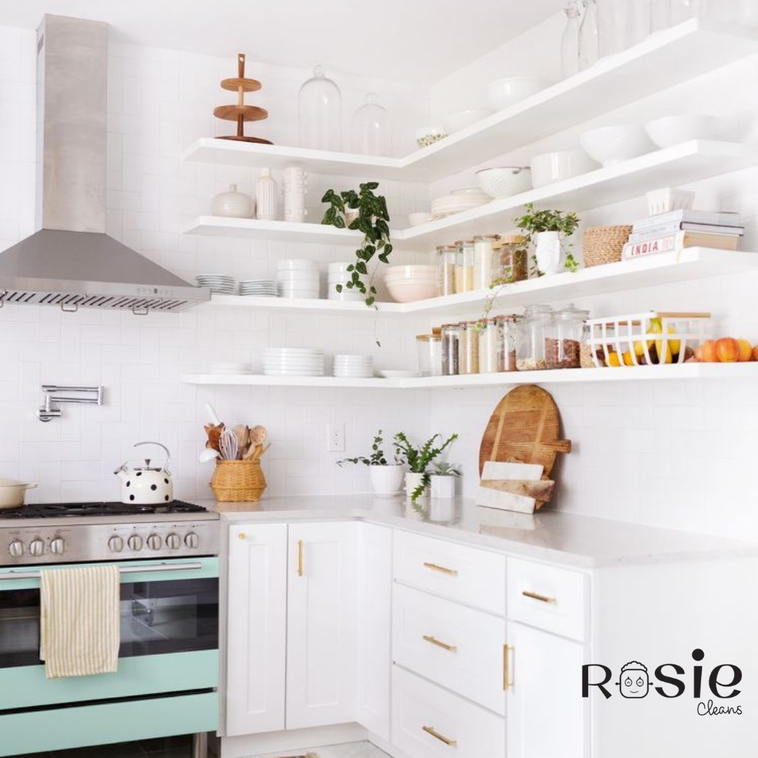 Bright white kitchen with open shelves holding dishes, bowls, jars, plants, and books, and a mint green oven with a white kettle.