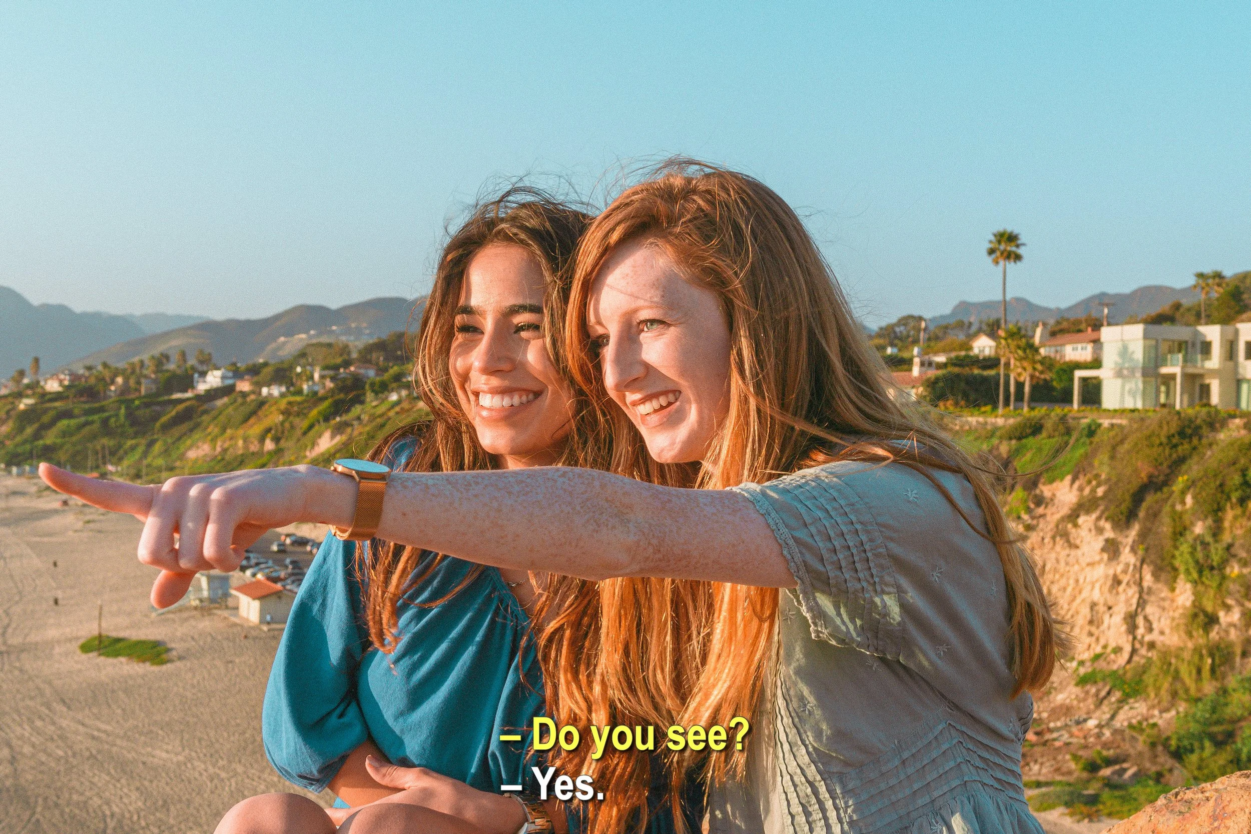 Two young women with long hair take a selfie on a beach with mountains, houses, and palm trees in the background. They are smiling and one of them is pointing at the camera.