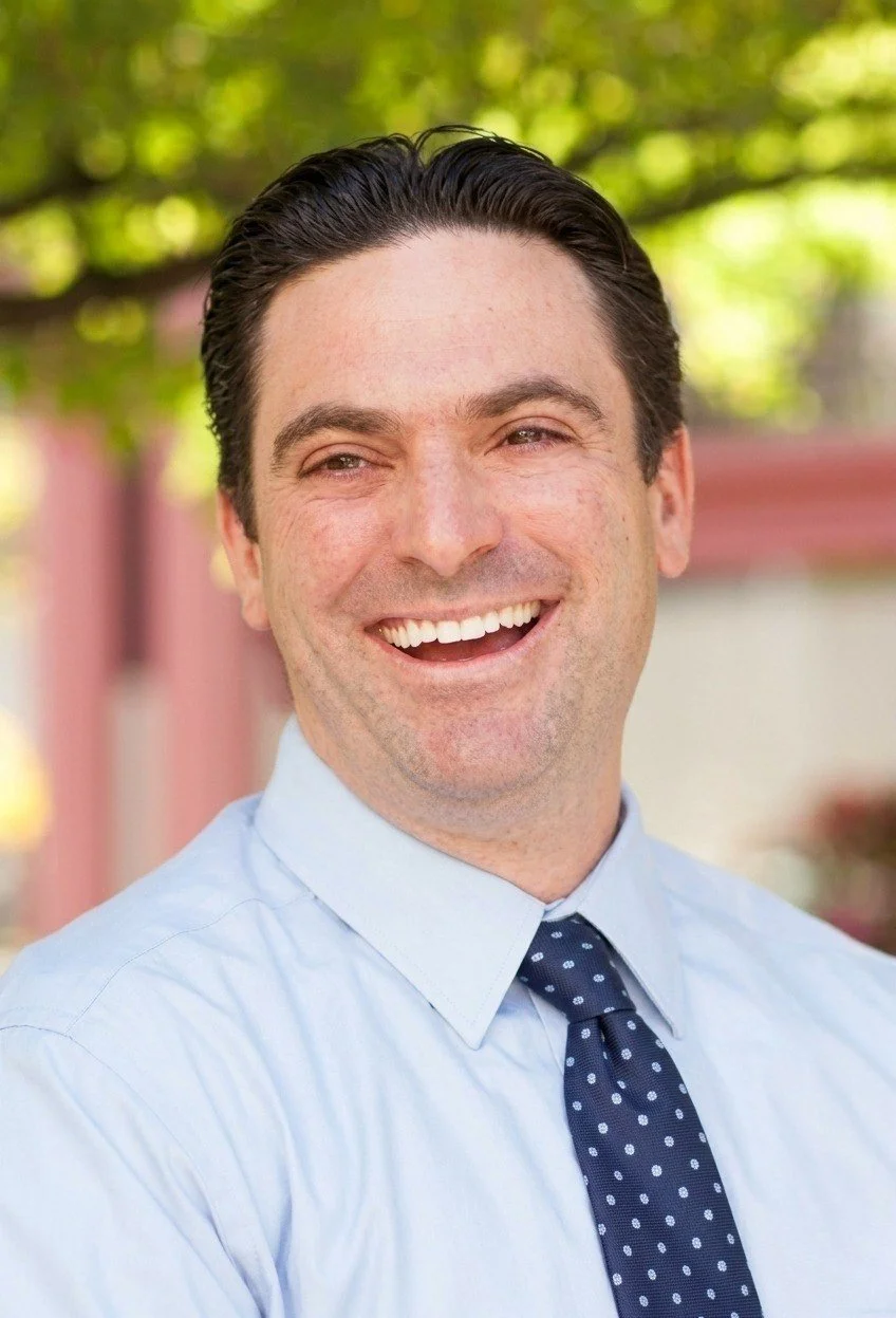 A man with dark hair, wearing a light blue shirt and navy blue tie with white polka dots, smiling outdoors with blurred green trees in the background.