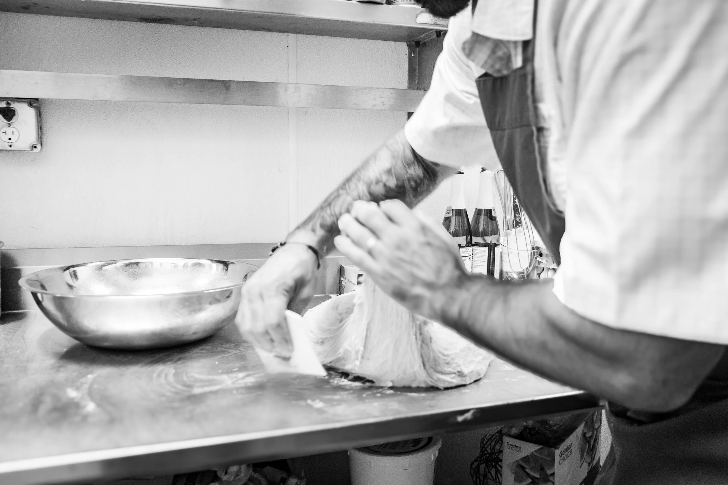 A person with tattoos on their arm is preparing meat on a kitchen counter. They are wearing a shirt and apron. There is a metal bowl, some condiments, and kitchen supplies in the background.