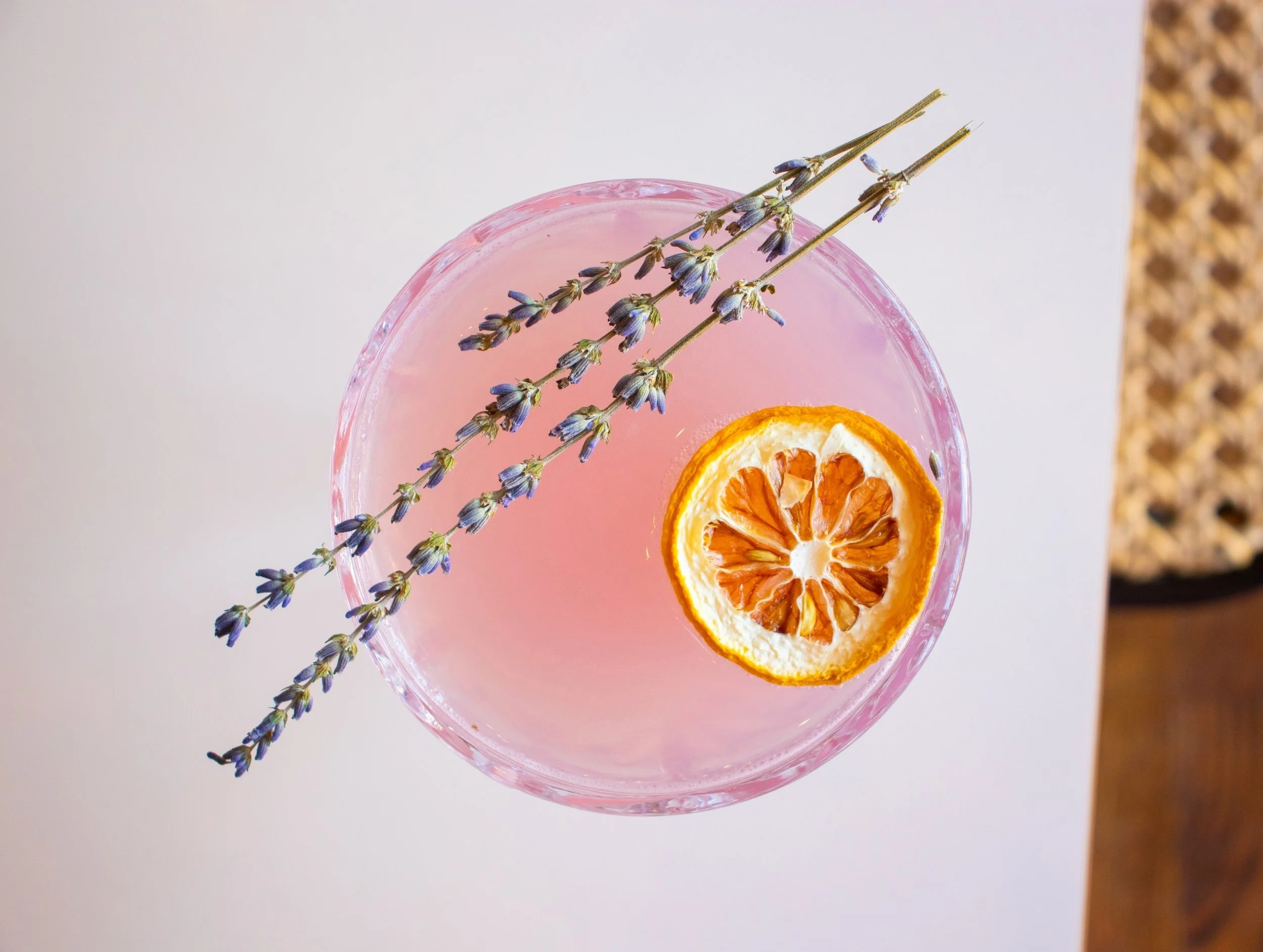 Pink cocktail in a glass garnished with dried orange slice and lavender sprigs.