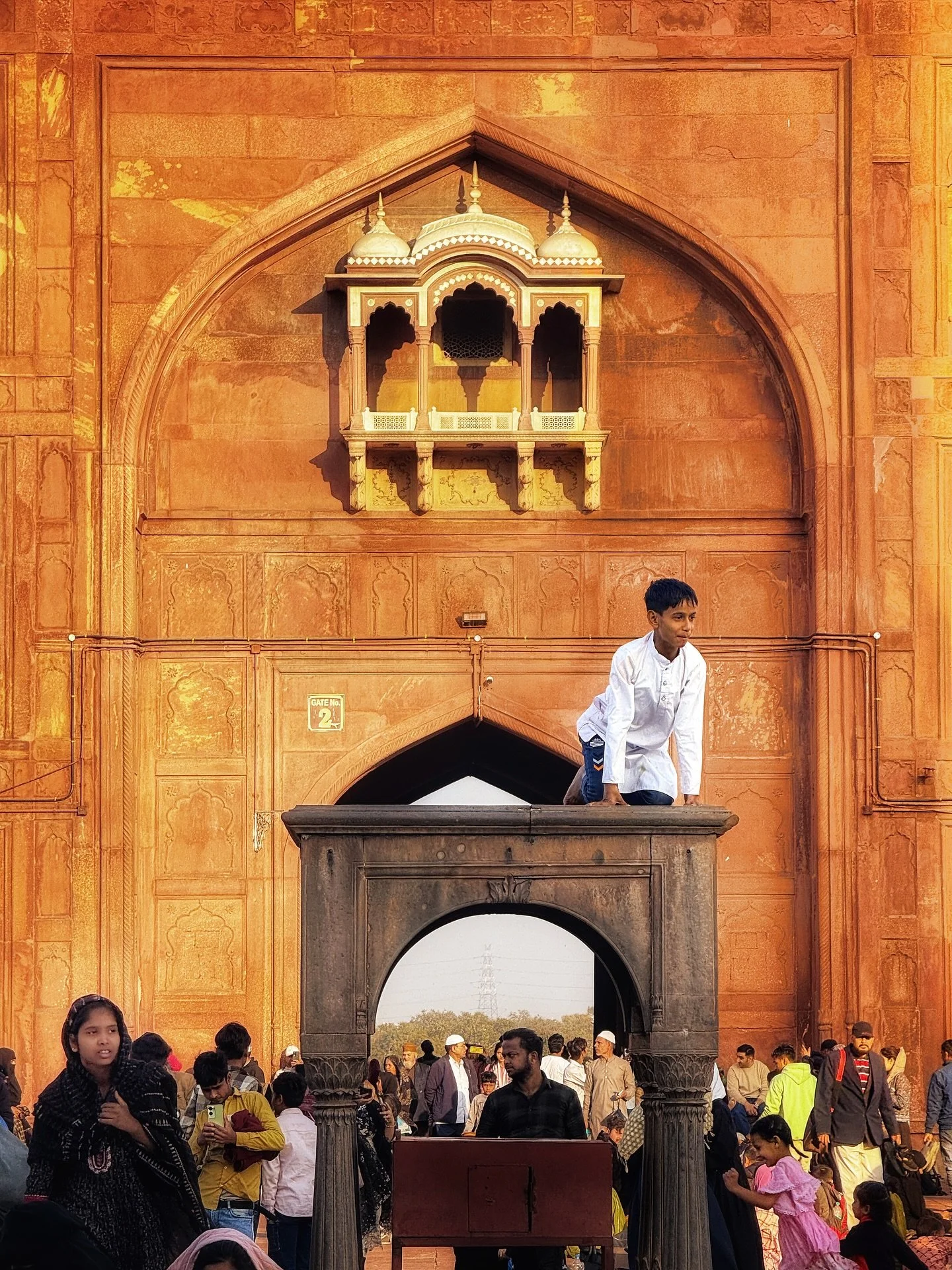 Part 2/3. I zoomed in a bit. And there he was: the little boy perched above the crowd, his white kurta catching the golden sun. He moved with the careless joy of childhood, the canopy became his throne, the crowd his court, the monument his kingdom.
