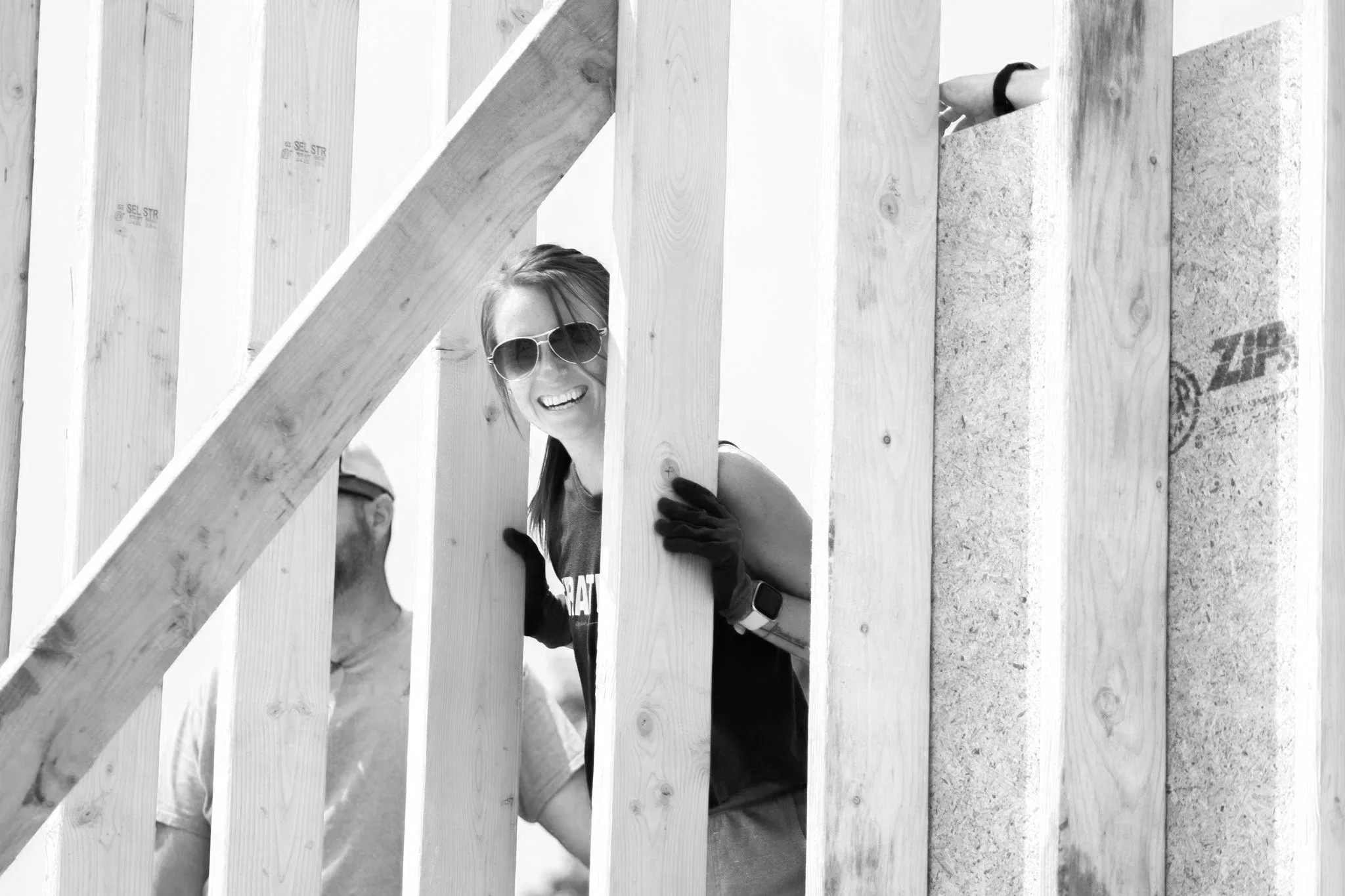 A woman with long hair, sunglasses, and a watch smiling and peering through the wooden frame of a building under construction.
