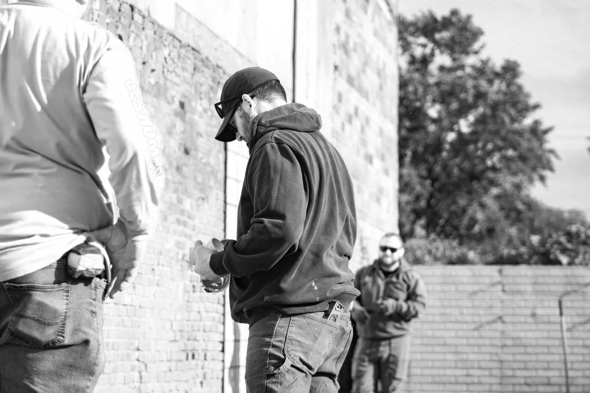 Three men outdoors near a brick wall, one with a handgun in his waistband, wearing a hoodie and cap, looking down at his phone; another in a jacket and cap, also on his phone; and a third in the background wearing sunglasses and a jacket.