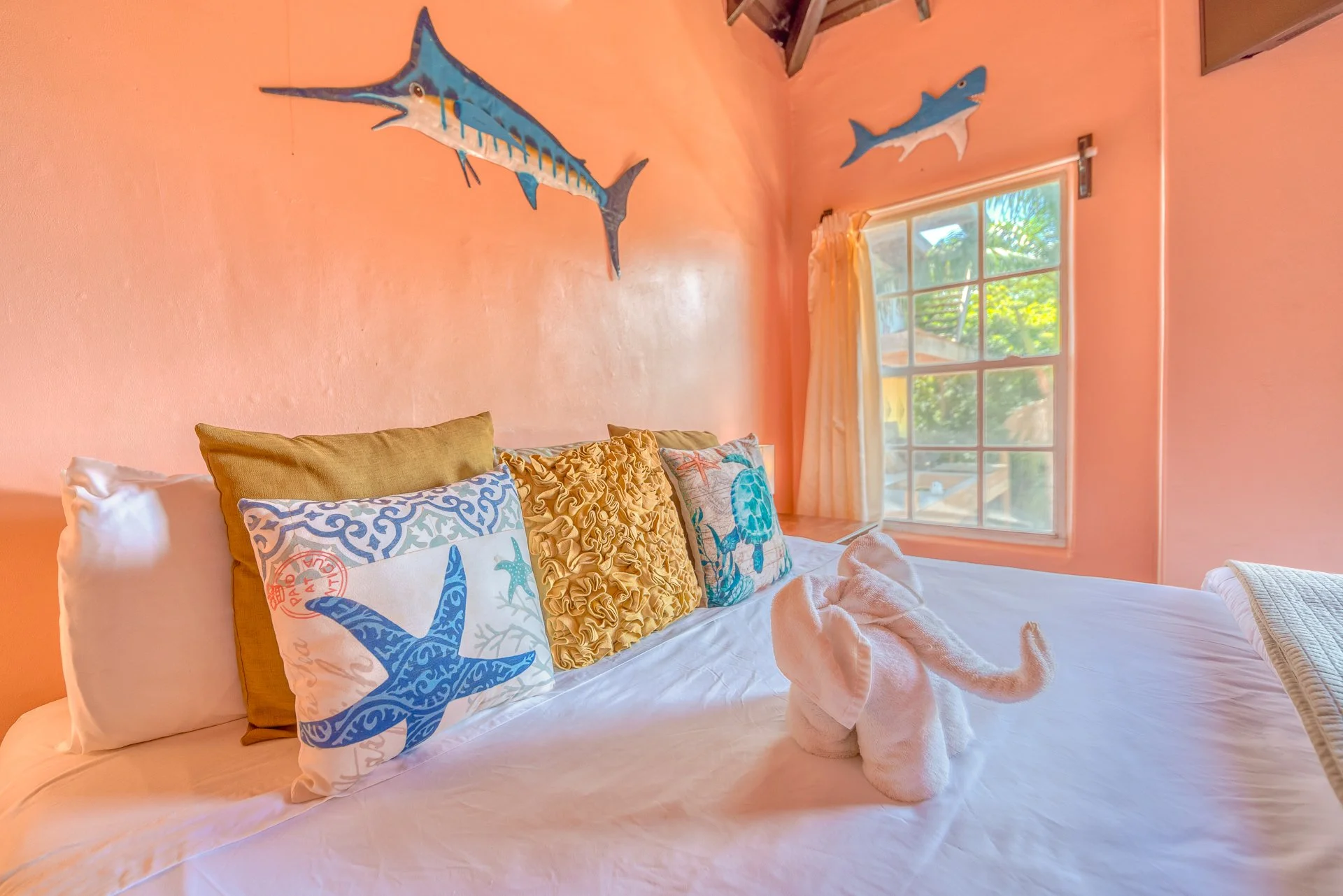 A bedroom with peach-colored walls decorated with mounted fish sculptures, a window with a view of greenery, and a bed with multiple decorative pillows, including one with a blue starfish design and another with ocean-themed prints.