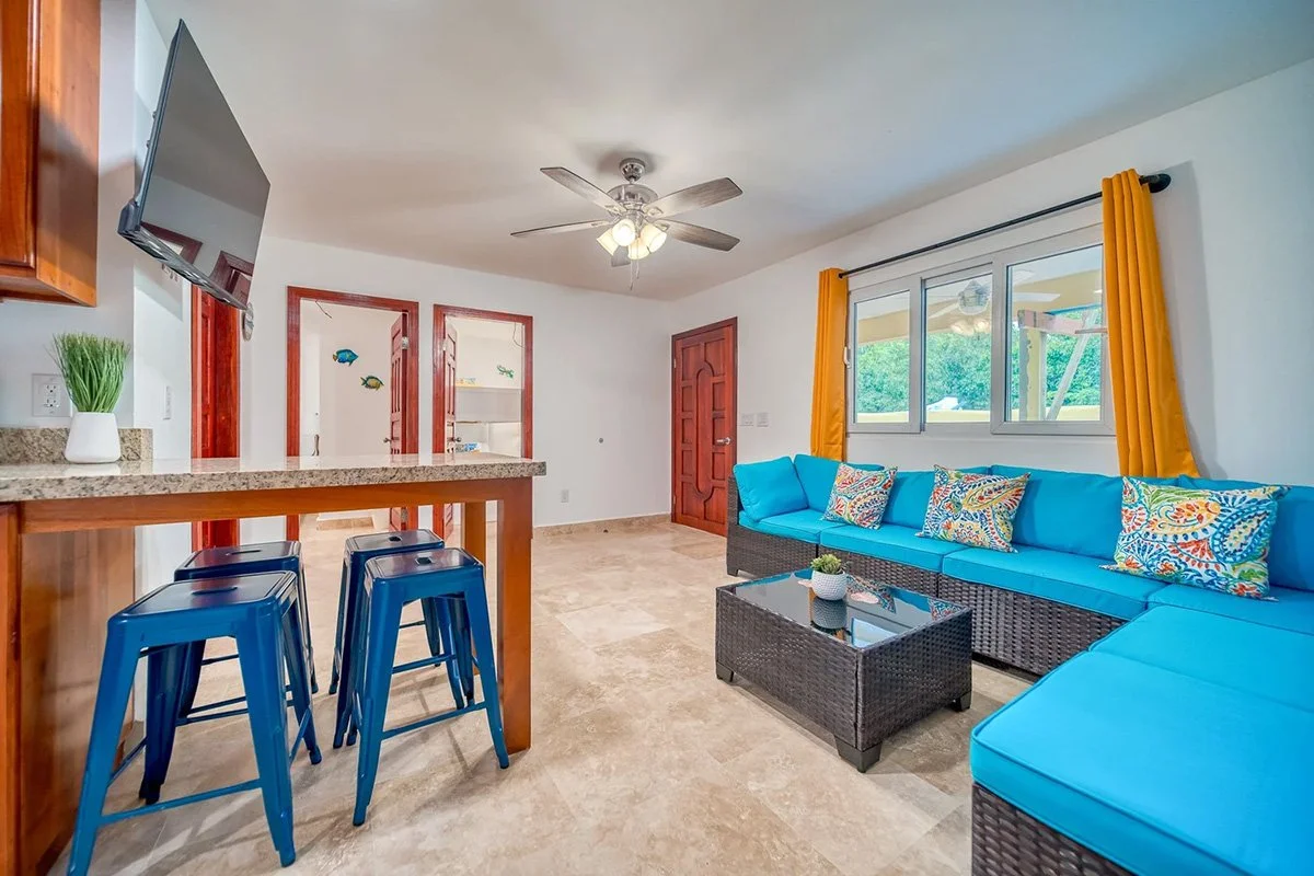 Living room with a blue sectional sofa, colorful pillows, a black wicker coffee table, a window with yellow curtains, a ceiling fan, and a wall-mounted TV.