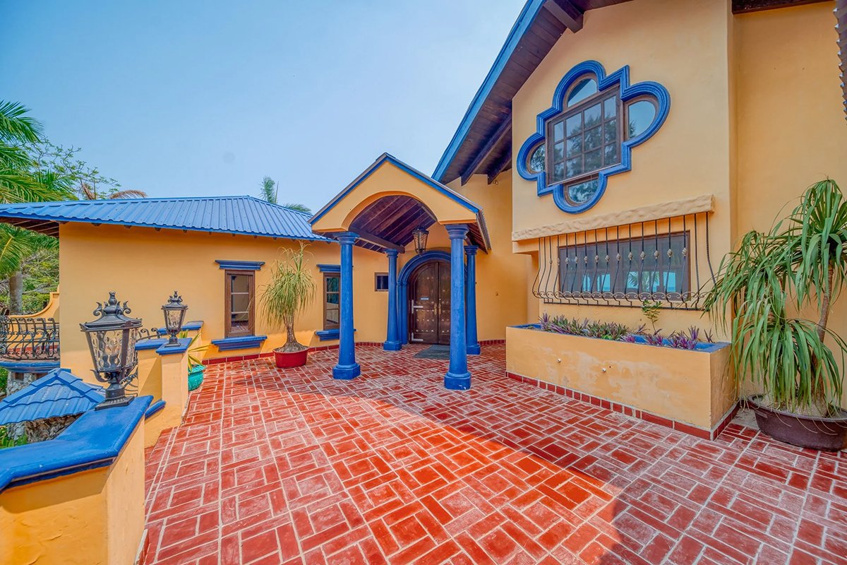 Colorful house with a yellow exterior, blue roof, and blue accents around the door and window. Red brick patio with decorative plants and lanterns.