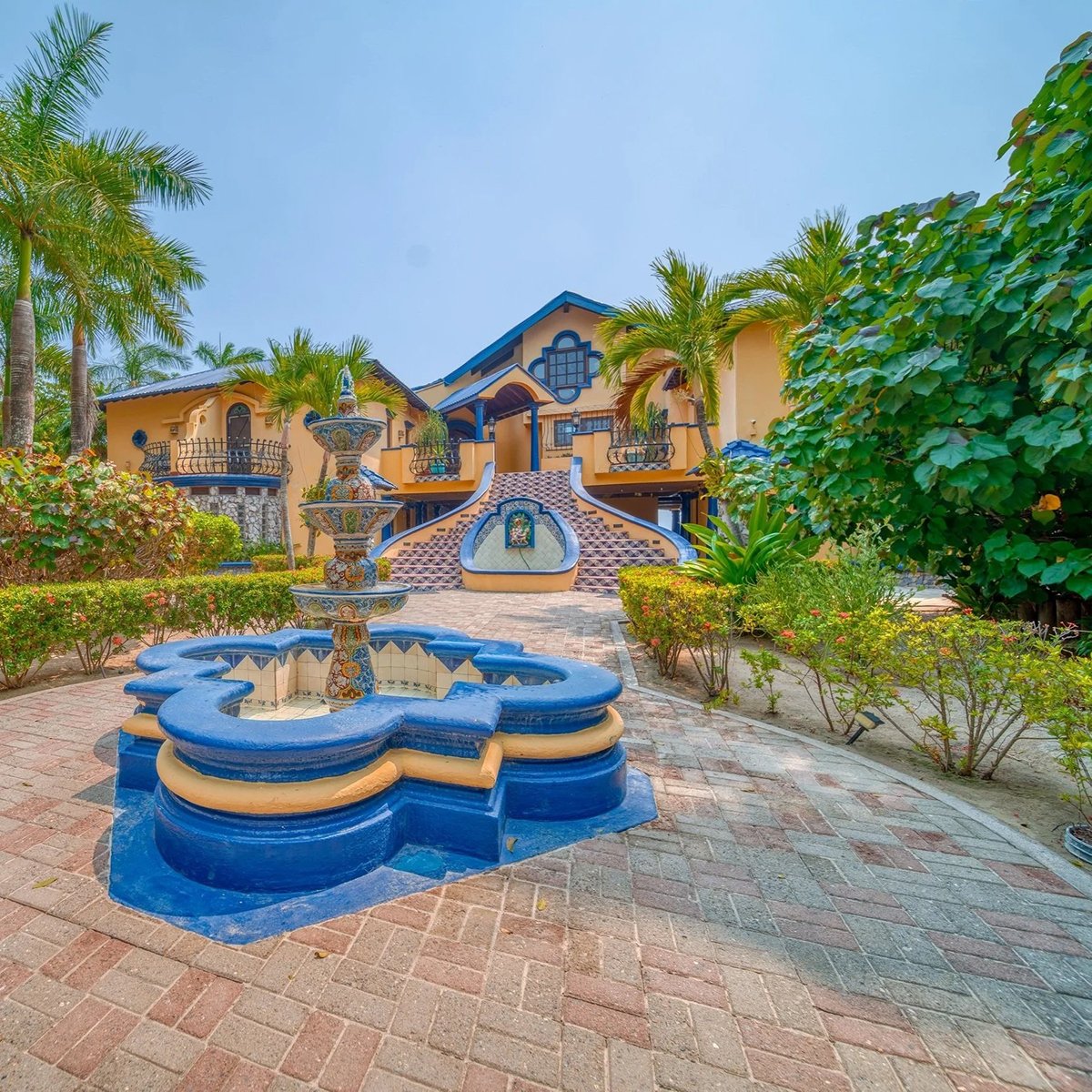 A colorful house with yellow walls and blue accents, surrounded by palm trees and greenery, featuring a decorative fountain in the foreground.