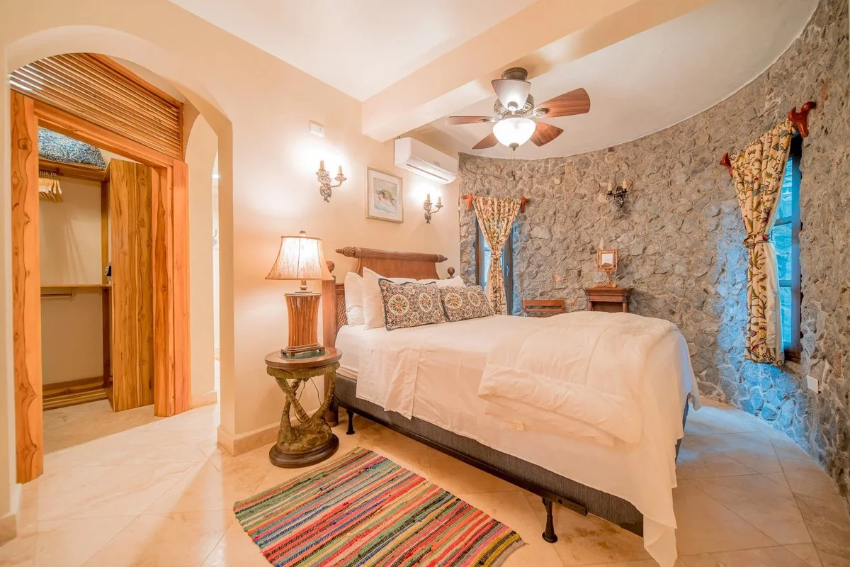 A bedroom with a stone wall, a wooden bed with white bedding and decorative pillows, floral curtains, a ceiling fan, a wooden nightstand, and a patterned rug on beige tile flooring.