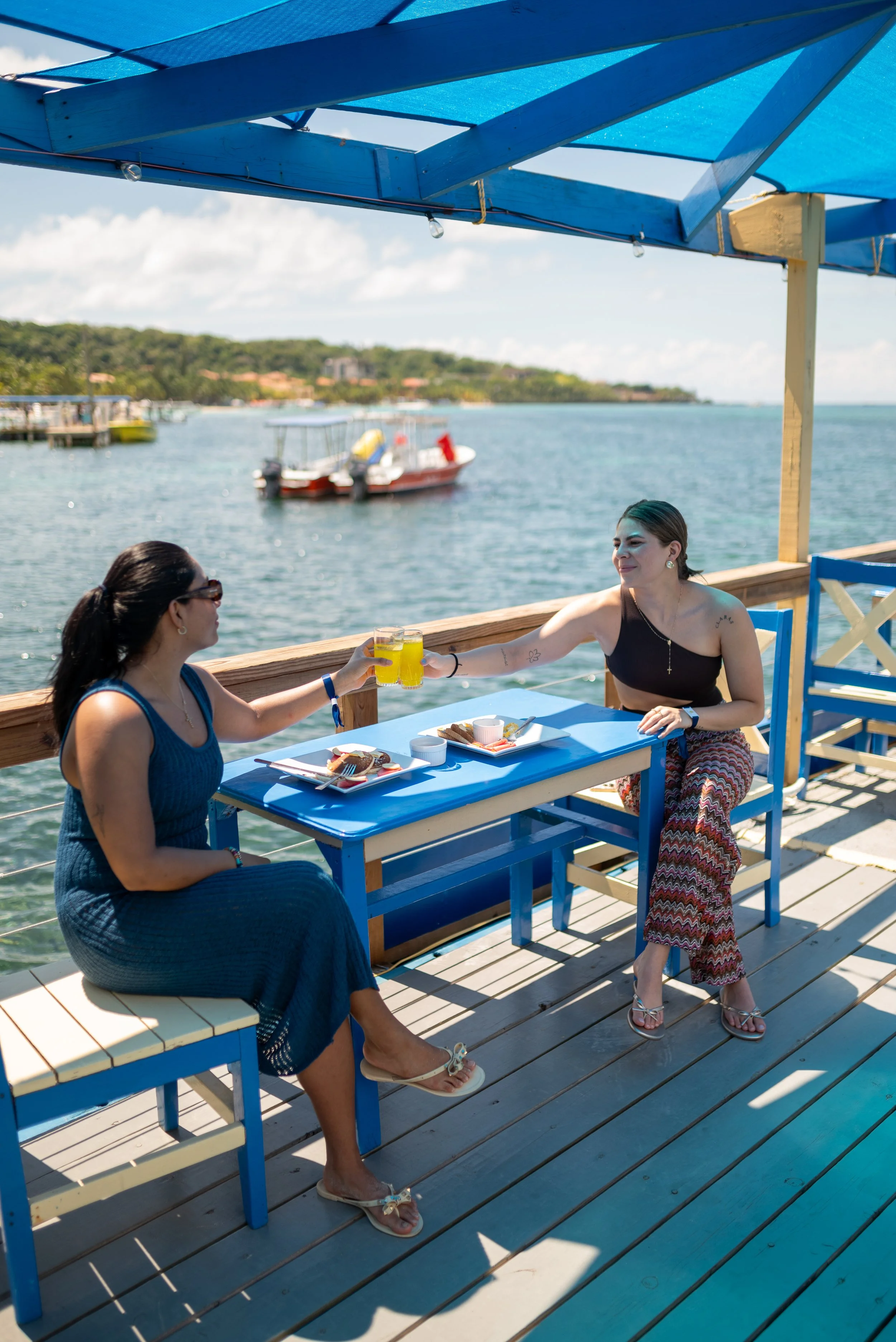 Two women sitting at an outdoor table near the water, raising glasses of yellow drinks for a toast, with a boat and water in the background.